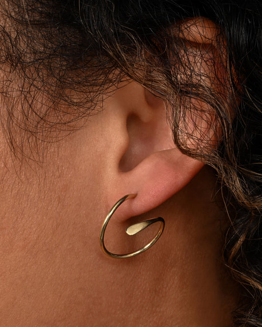 A yellow gold earring in a loop shape with one hammered flat end. Shown on a person from the front.
