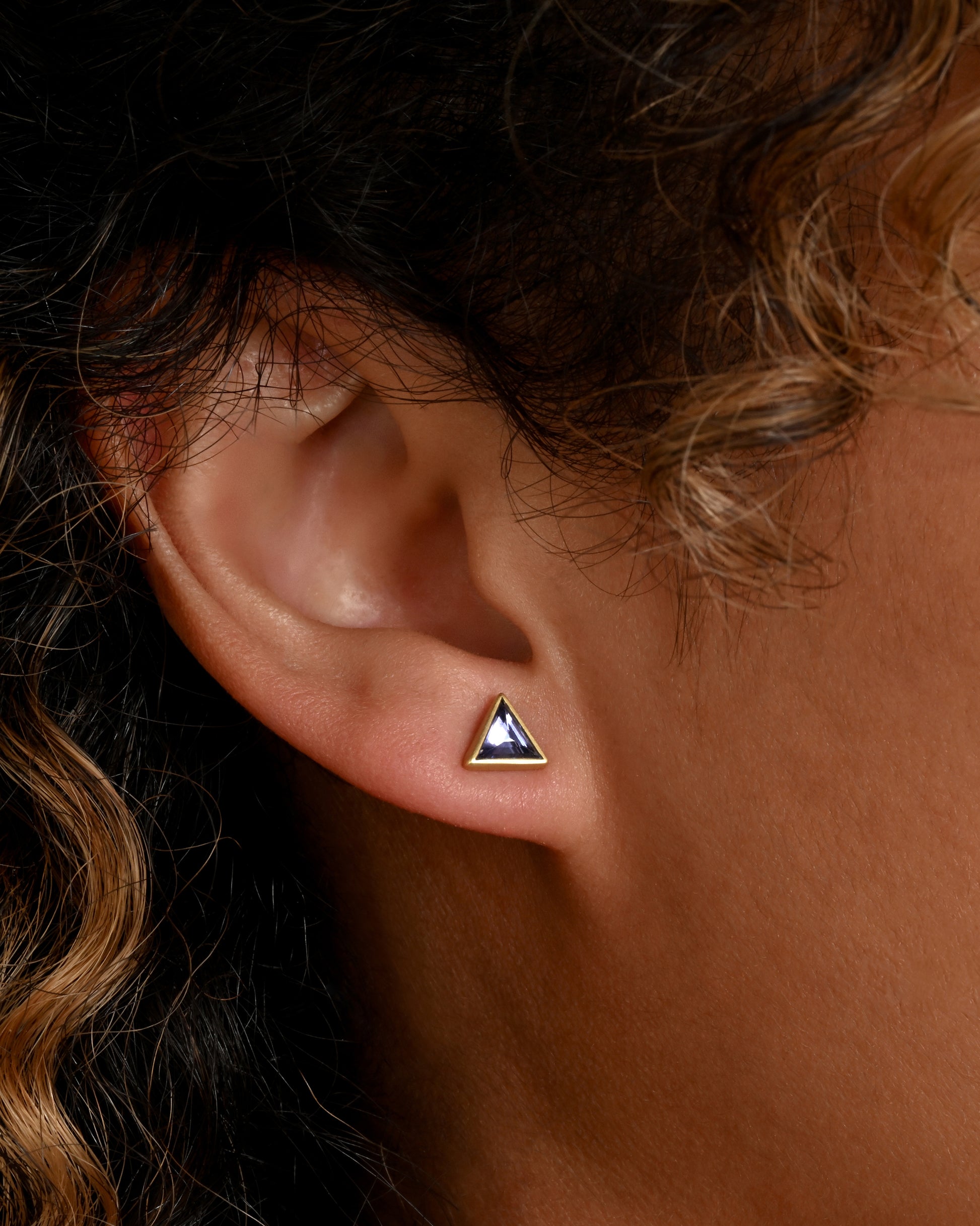 A pair of yellow gold triangle studs with pale blue bezel set sapphires. Shown on an ear.