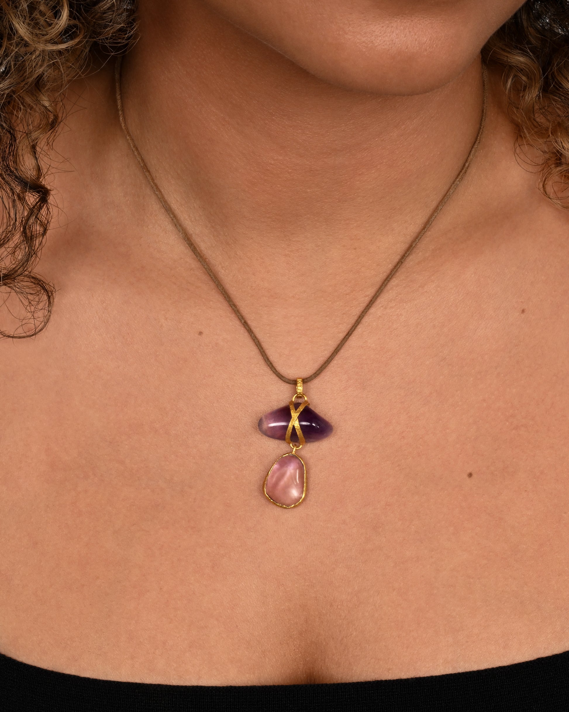 An amethyst pendant wrapped in high karat yellow gold hanging from a cord necklace. Shown worn around the neck.