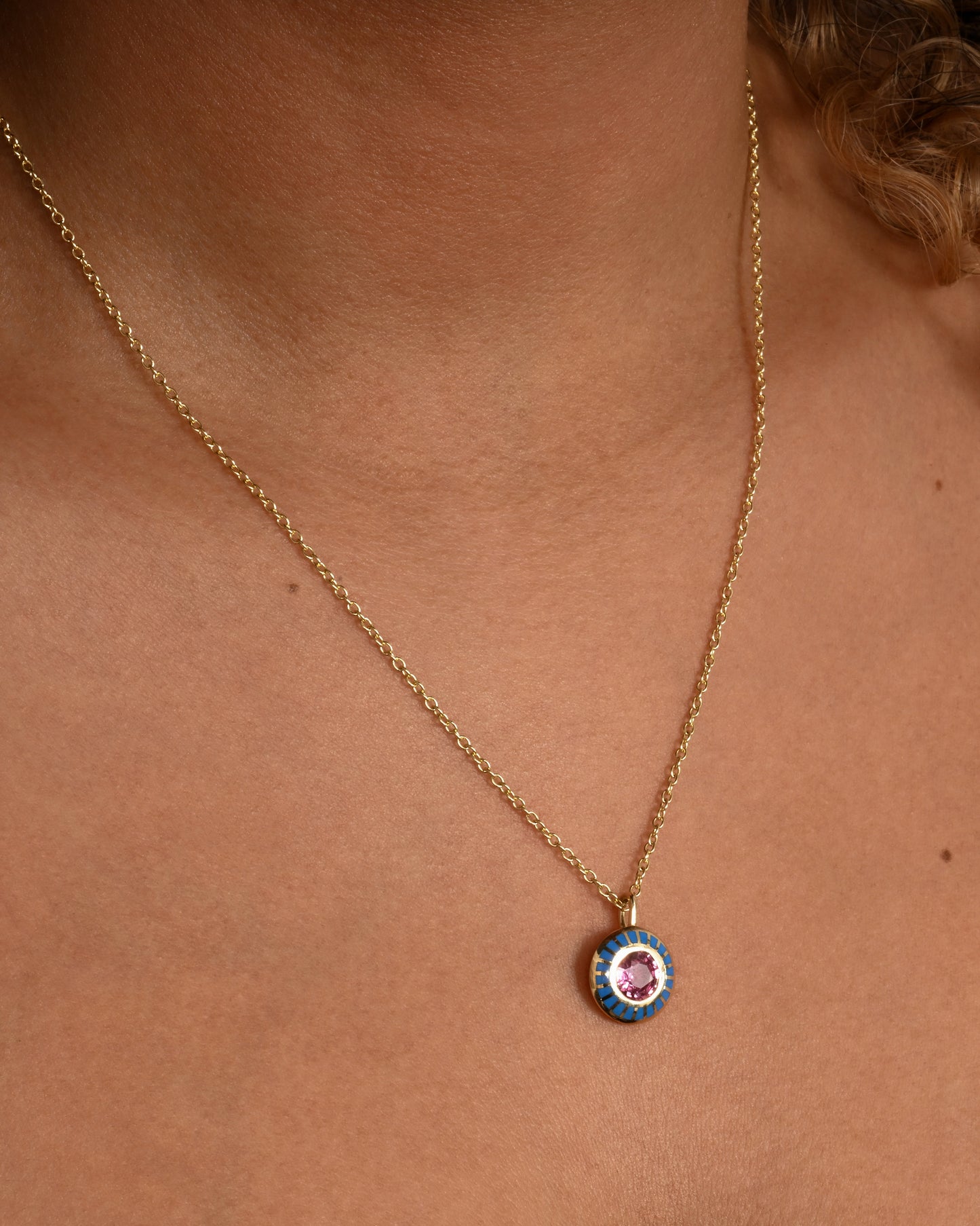 A yellow gold pendant with light blue enamel surrounding a pink sapphire. Shown on a cable chain, worn.