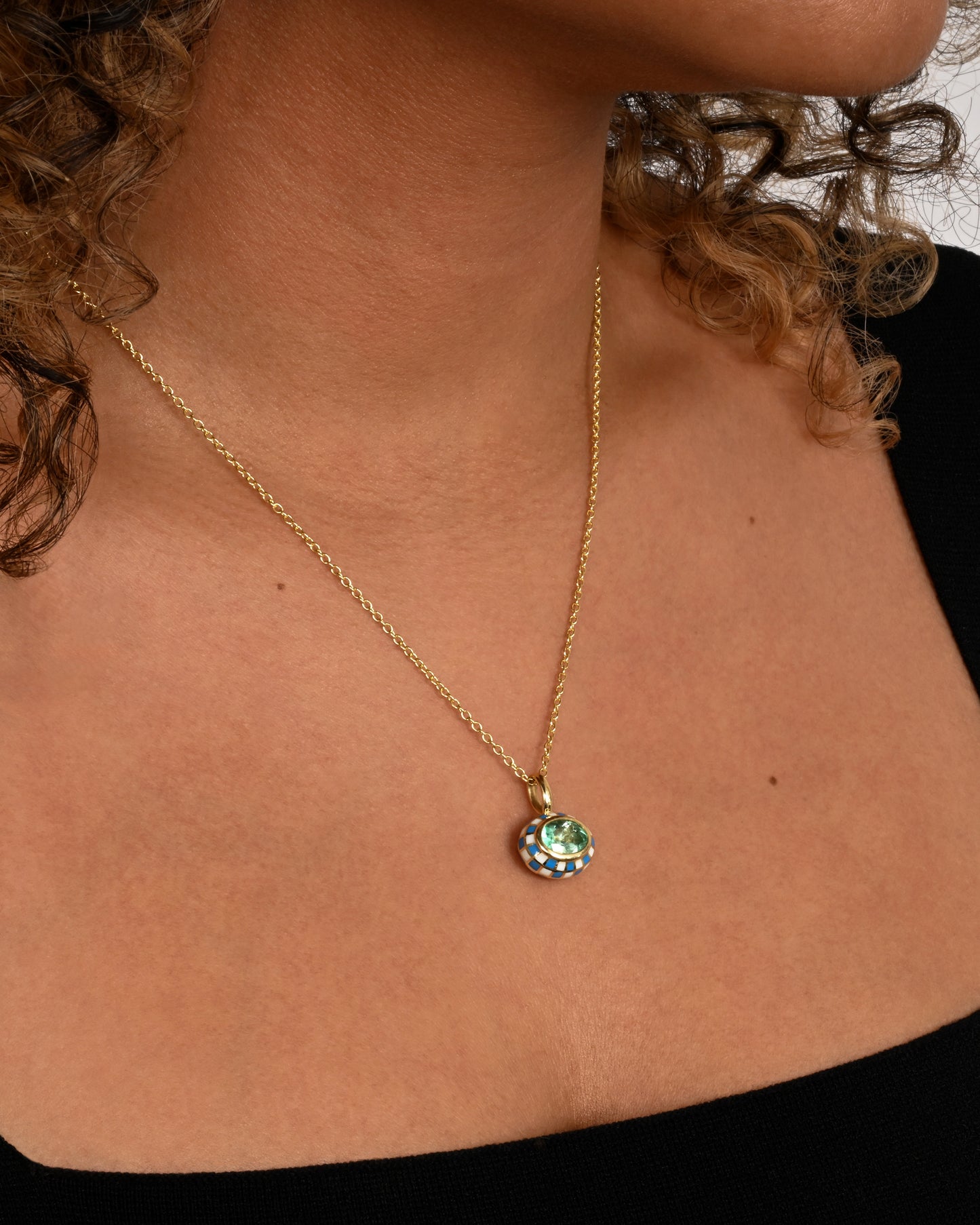 A yellow gold oval pendant with blue and white enamel surrounding an emerald. Shown hanging from a cable chain worn.