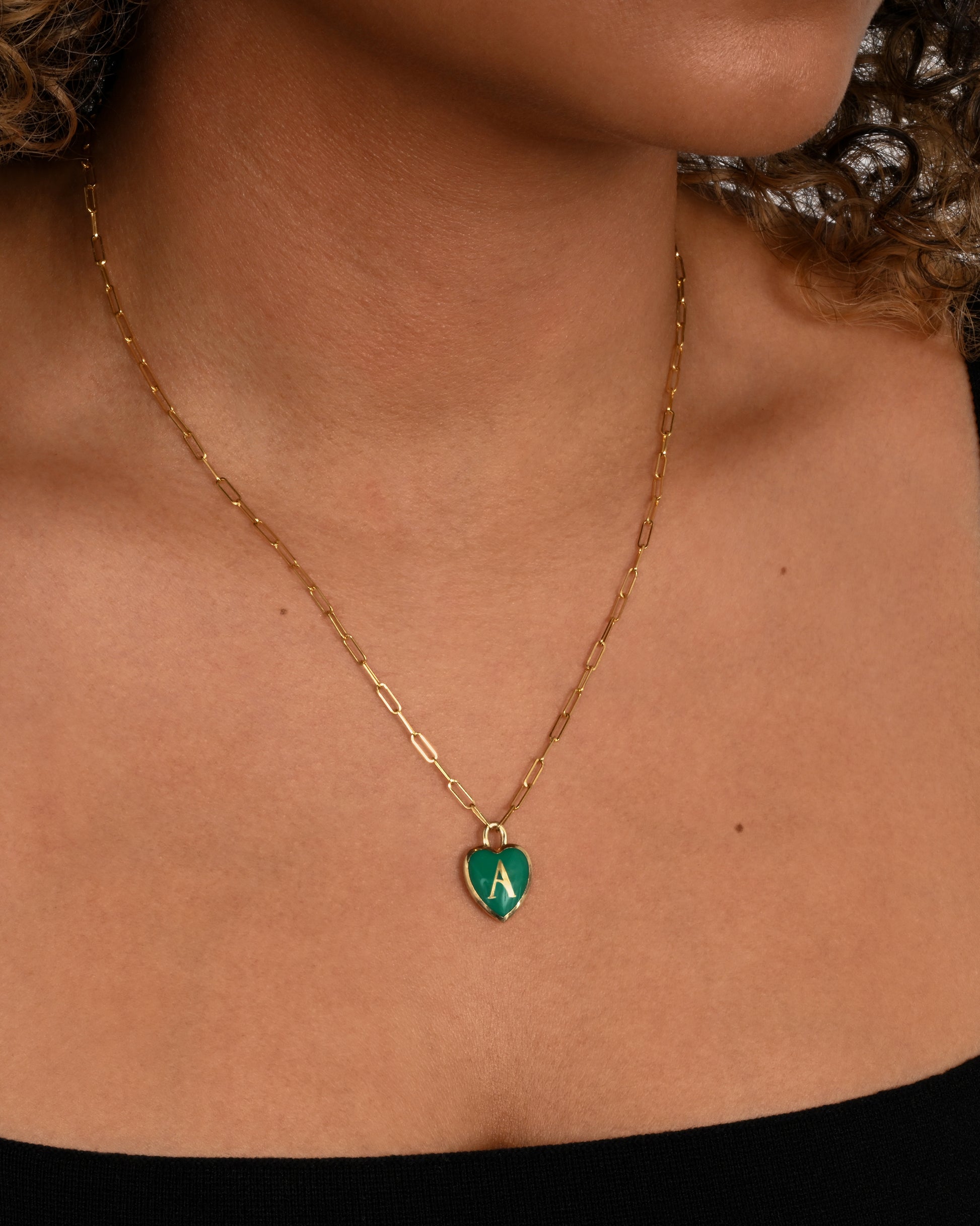 A yellow gold paper clip chain necklace with a heart shaped pendant that has green enamel and the letter "A". Shown around a neck.