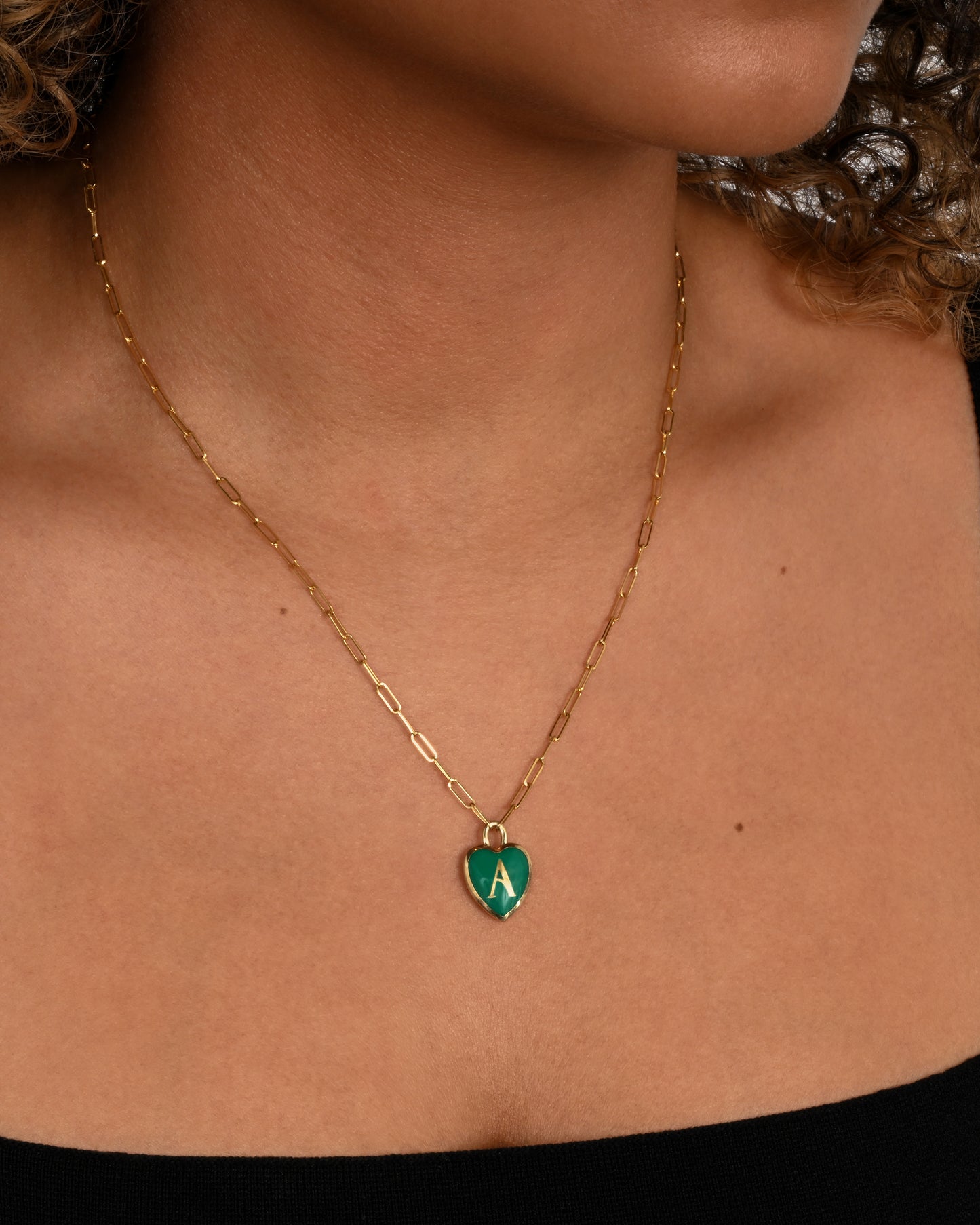 A yellow gold  paperclip chain necklace with a green enamel heart shaped A pendant. Shown from the front on a person.