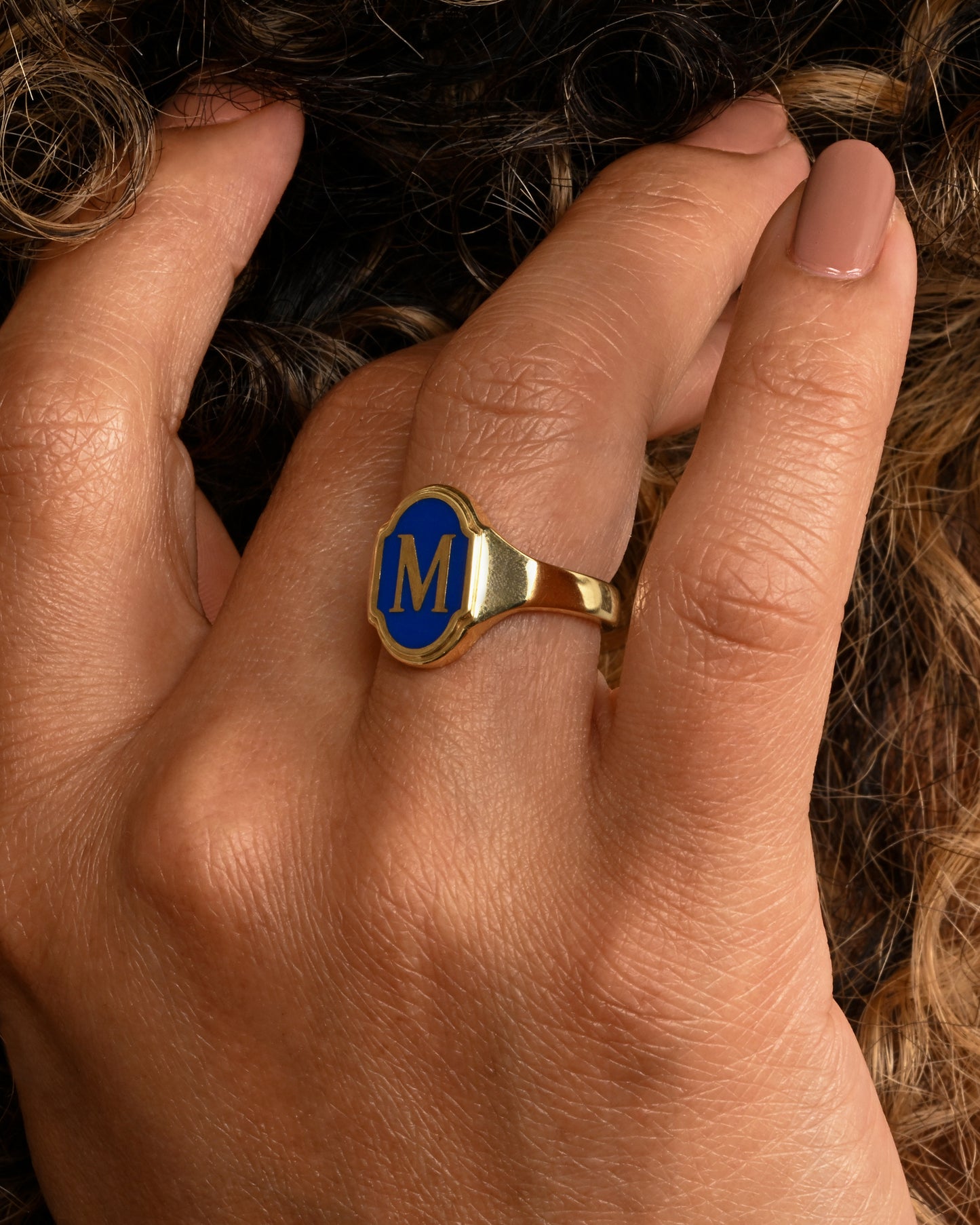 A yellow gold signet ring with blue enamel and the letter "M". Shown on a hand.