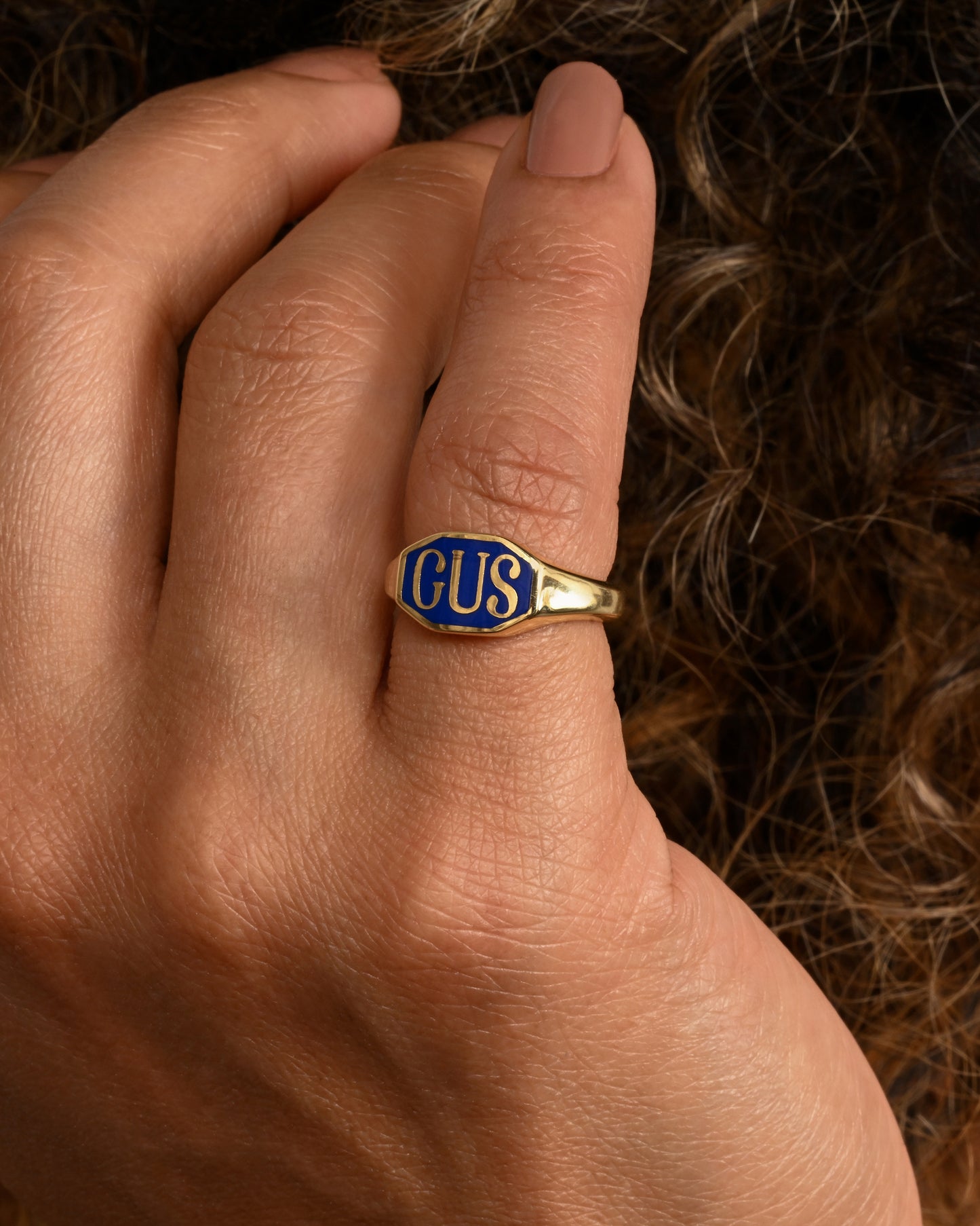 A yellow gold signet ring with the initials "GUS" and blue enamel. Shown worn on the pinkie finger.