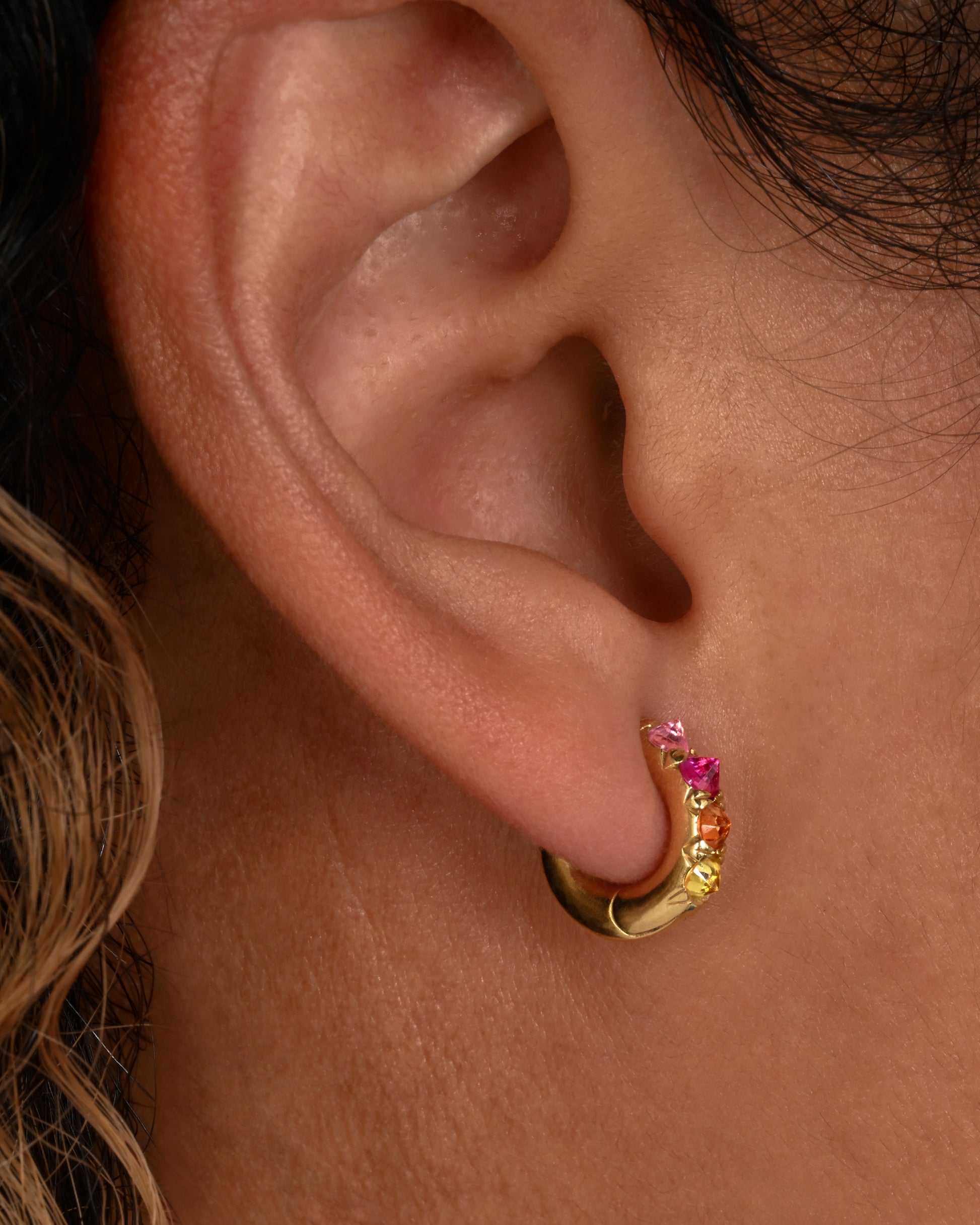 Gradient pink and orange inverted sapphires set in 18k yellow gold huggie hoops. Shown on an ear.