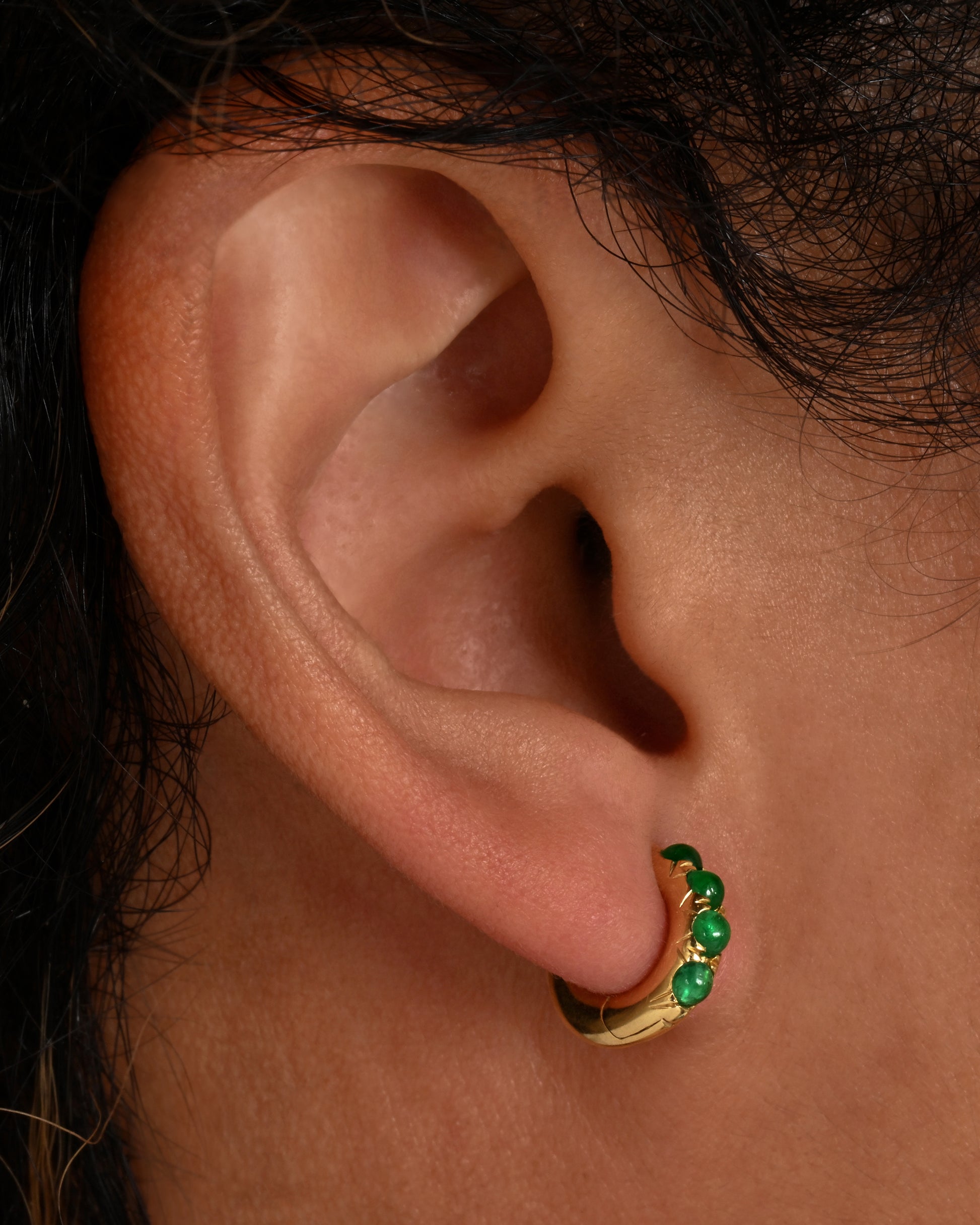 A pair of chunky huggie hoops with cabochon emeralds. Shown on an ear.