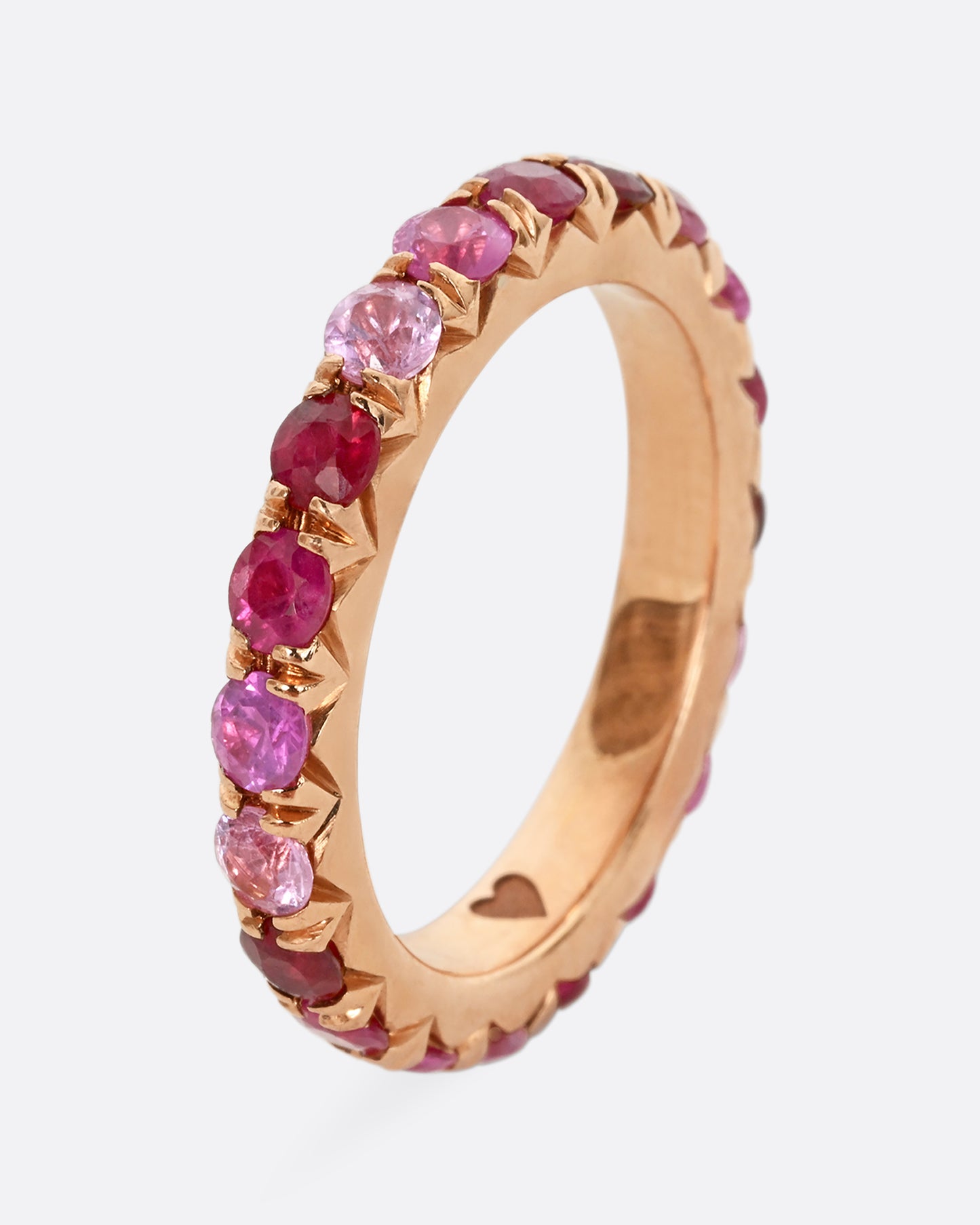 A rose gold eternity band with pink and red cut rubies. Shown from the side, standing up.