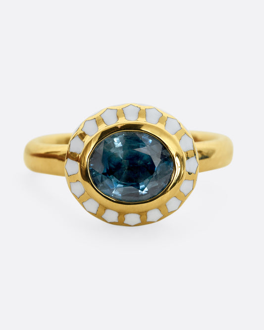 A yellow gold ring with a light blue sapphire and white enamel halo. Shown from the front, close up.