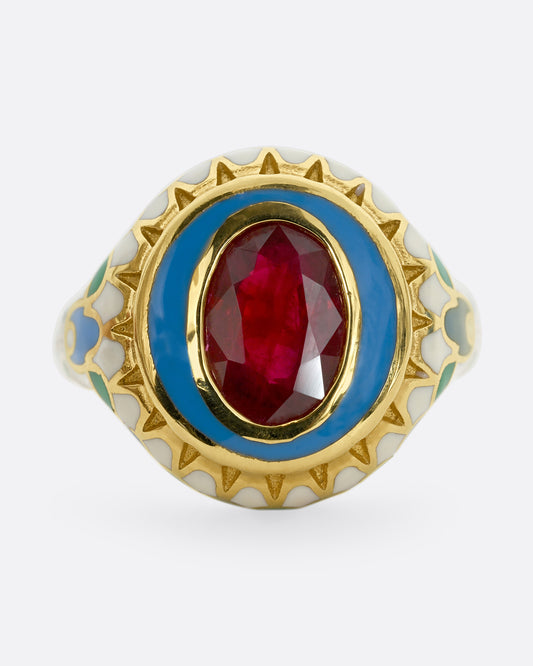 A yellow gold ring with multicolor enamel around an oval shaped ruby. Shown from the front.