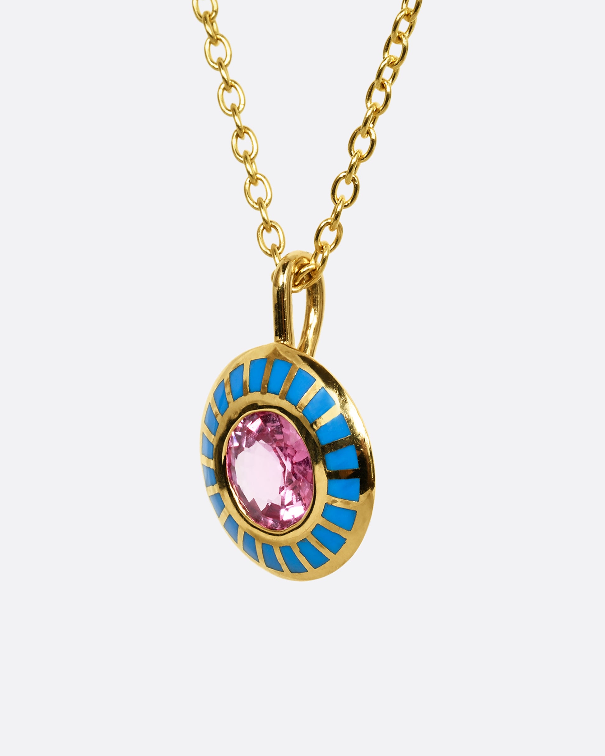 A yellow gold pendant with light blue enamel surrounding a pink sapphire. Shown on a cable chain from the side, close up.