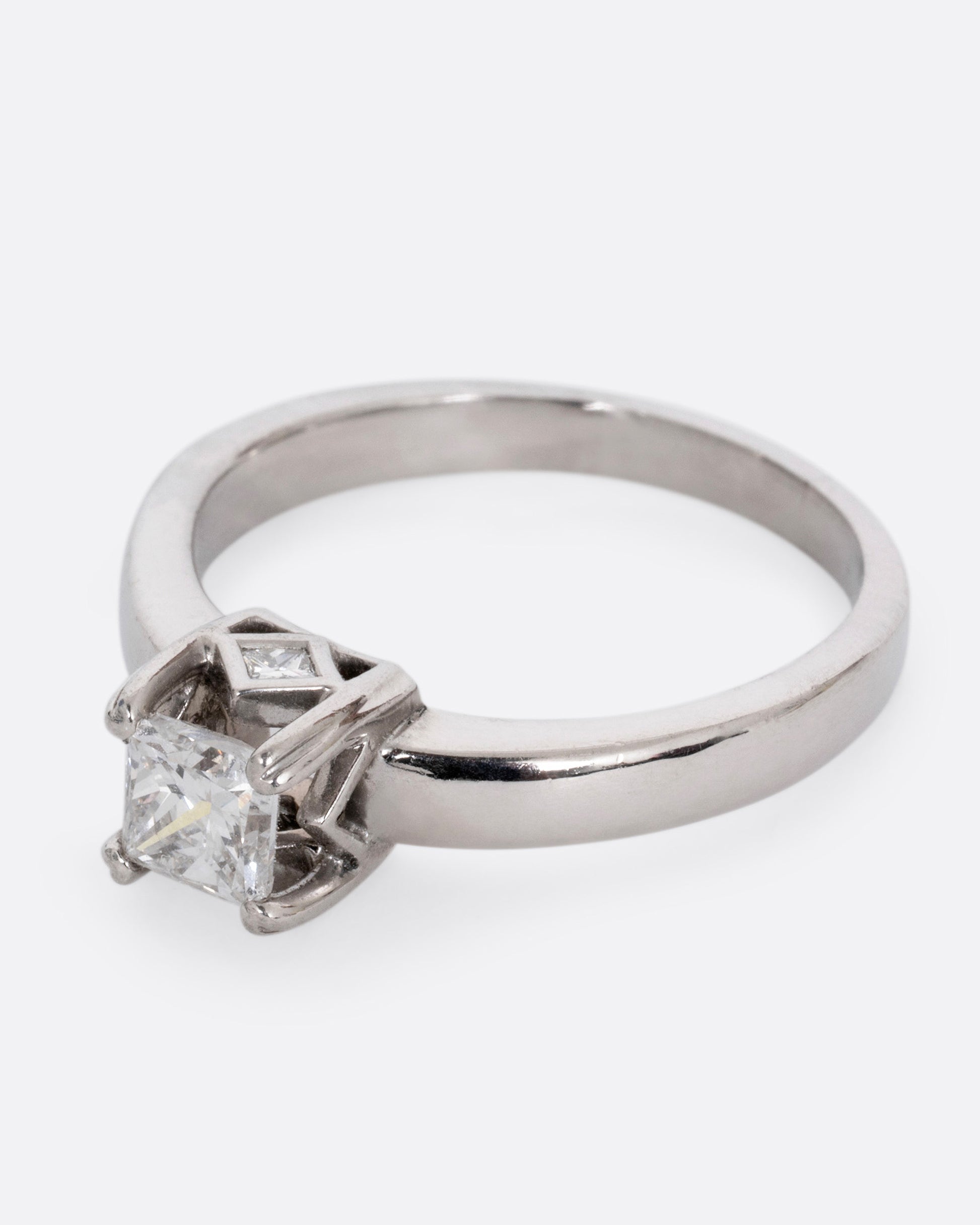 A platinum ring with a princess cut diamond, prong set high above angular cutouts and diamond accents. Shown from the side.