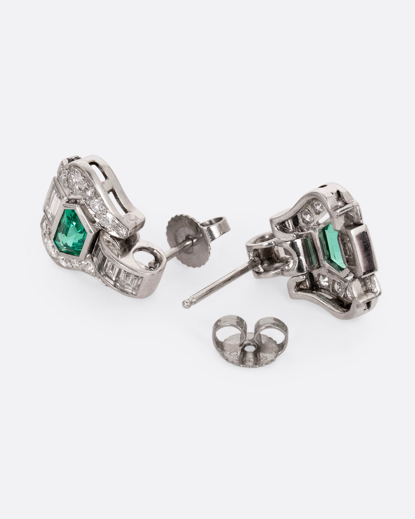 A pair of platinum earrings with emeralds and diamonds, shown lying on their sides, one from the front, the other from the back open.