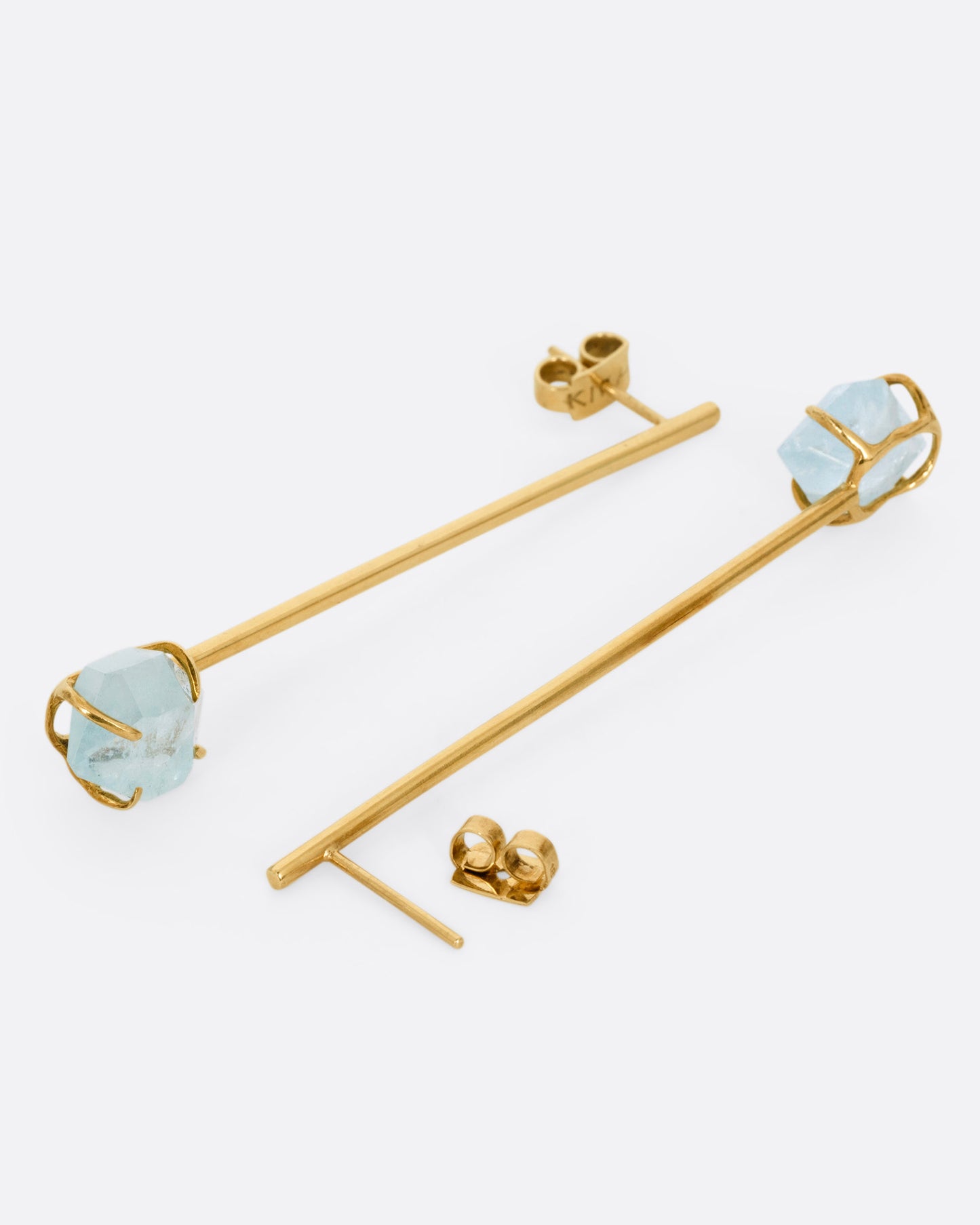 A pair of yellow gold bar earrings with  prong set uncut aquamarines at the base, shown lying on their sides with one earring open.