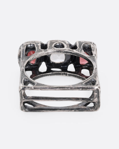 A silver ring with a cushion cut white topaz between two half moon garnets, shown from the back.