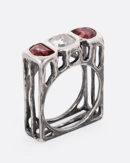 A silver ring with a cushion cut white topaz between two half moon garnets, shown standing up from the side.