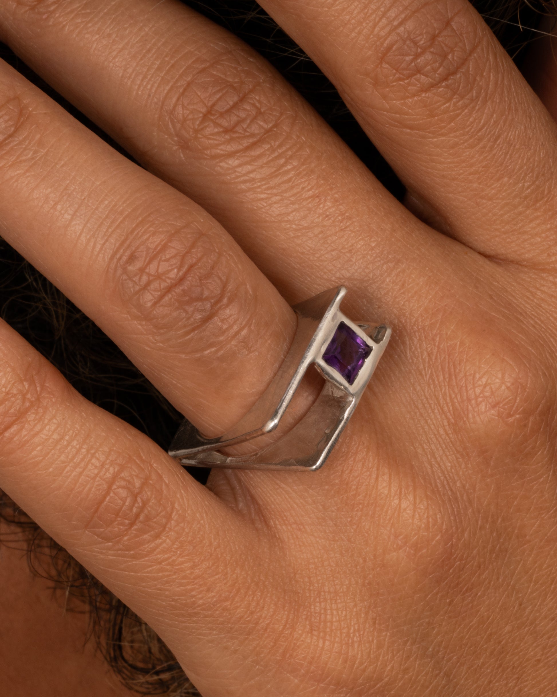 A silver square ring that widens to reveal a princess cut amethyst, shown on a hand.