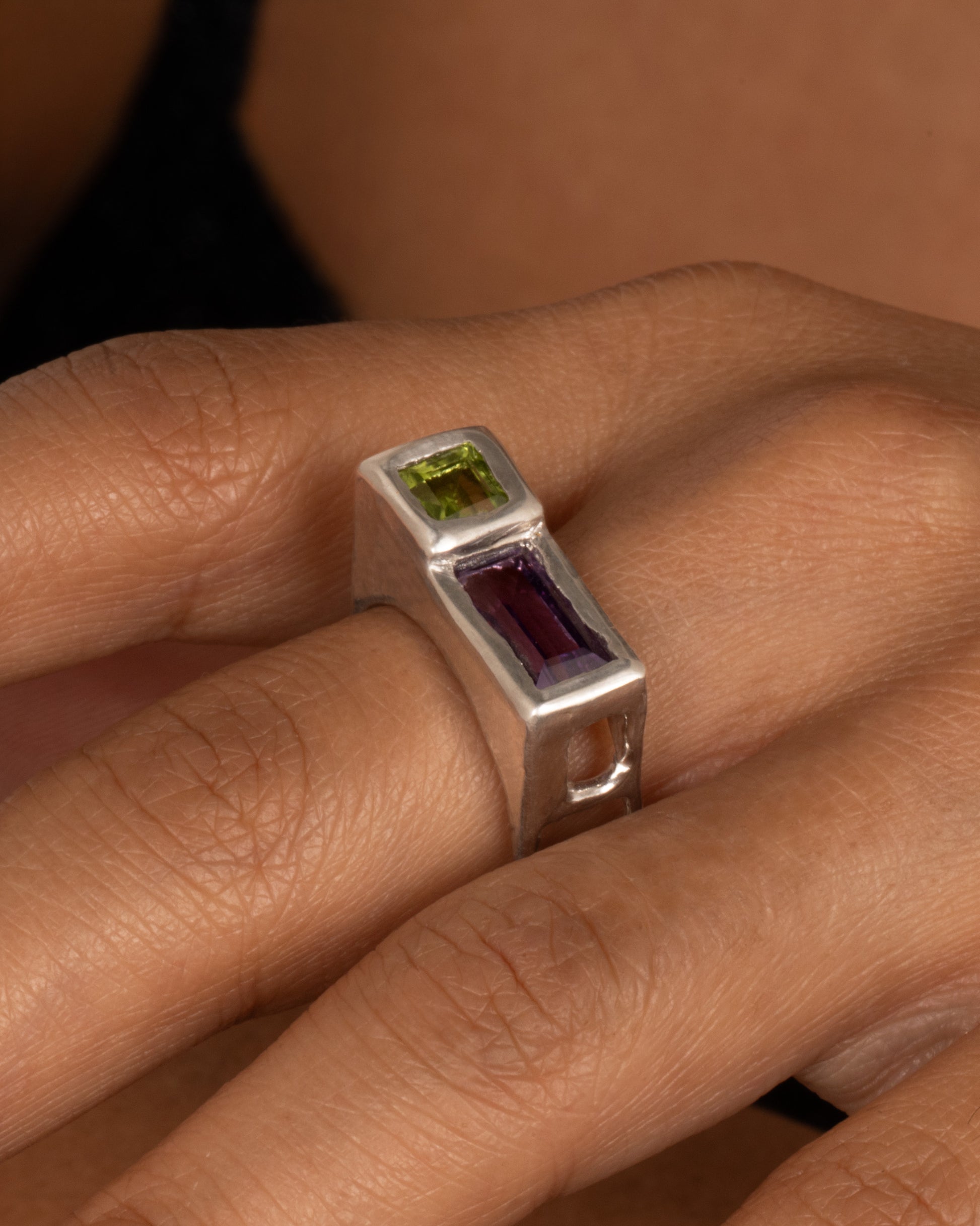 A silver square ring with a princess cut peridot and baguette amethyst, shown on a hand.
