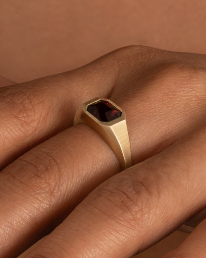 A yellow gold signet ring with a garnet, shown on a hand.