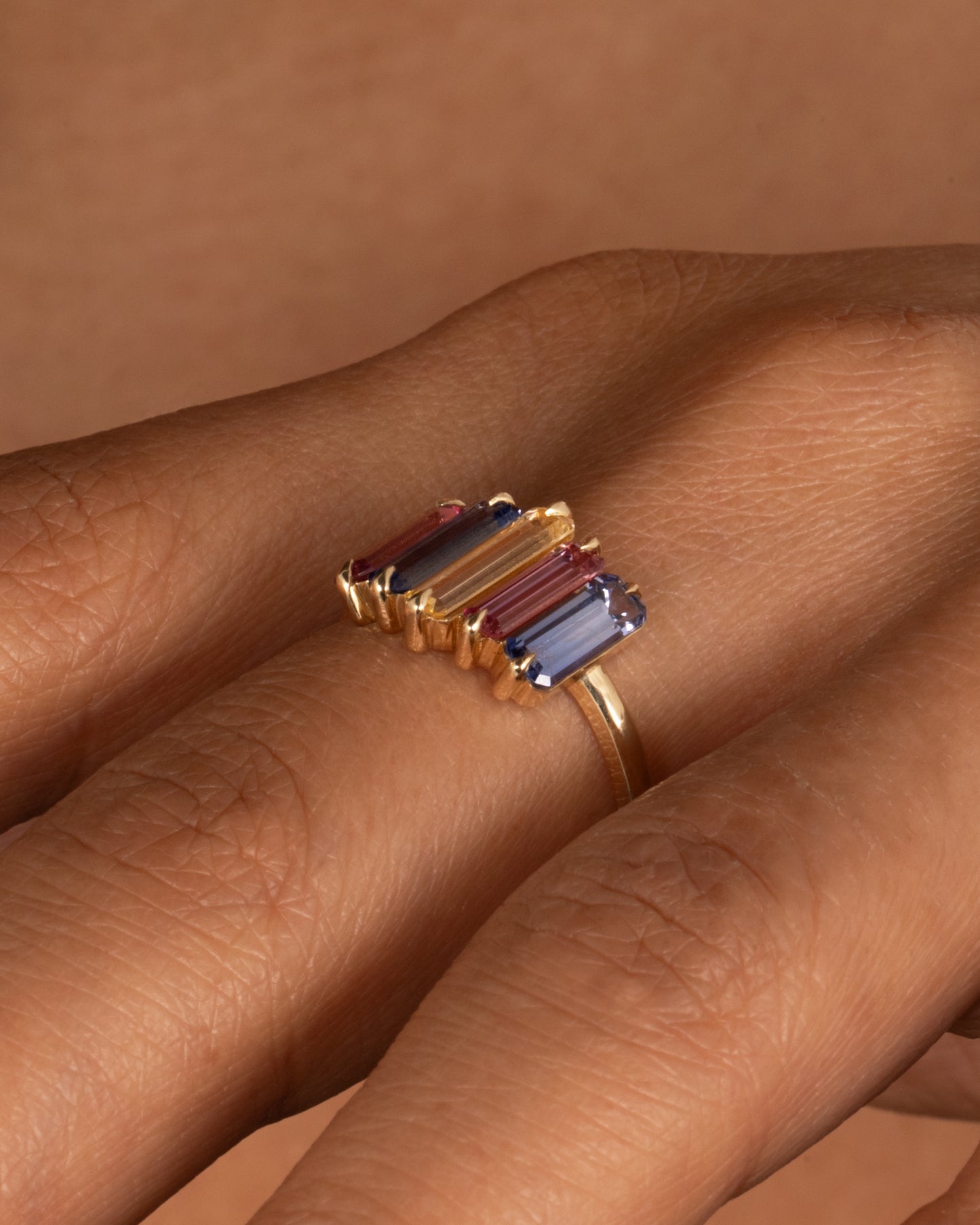 A yellow gold queue of pastel colored sapphire baguettes, shown on a hand.