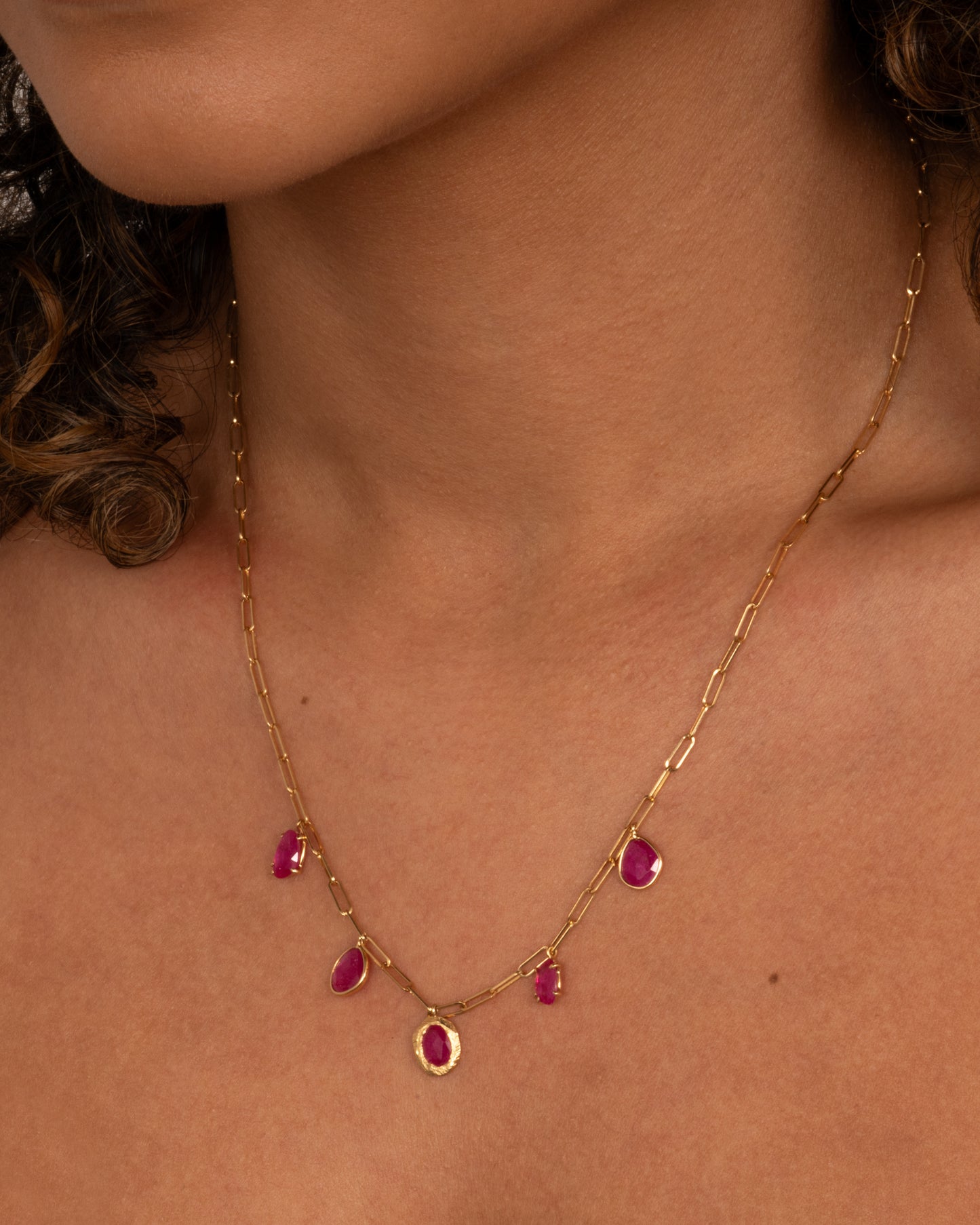 A yellow gold staple chain necklace with five rose cut ruby charms. Shown worn around the neck.