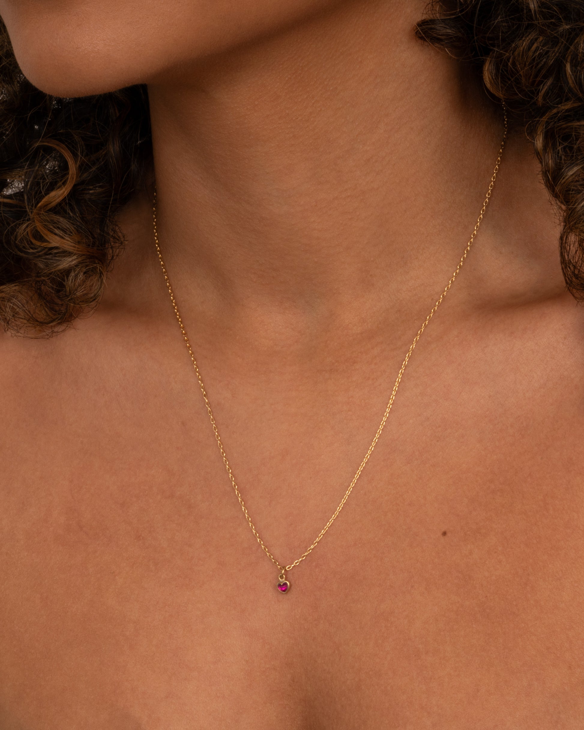 A yellow gold cable chain necklace with a tiny dimensional ruby heart, shown from the front on a person.