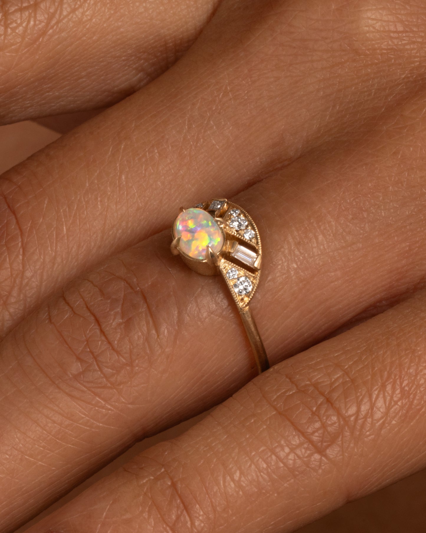 A yellow gold ring with a prong set opal and a diamond studded halo, shown on a hand.