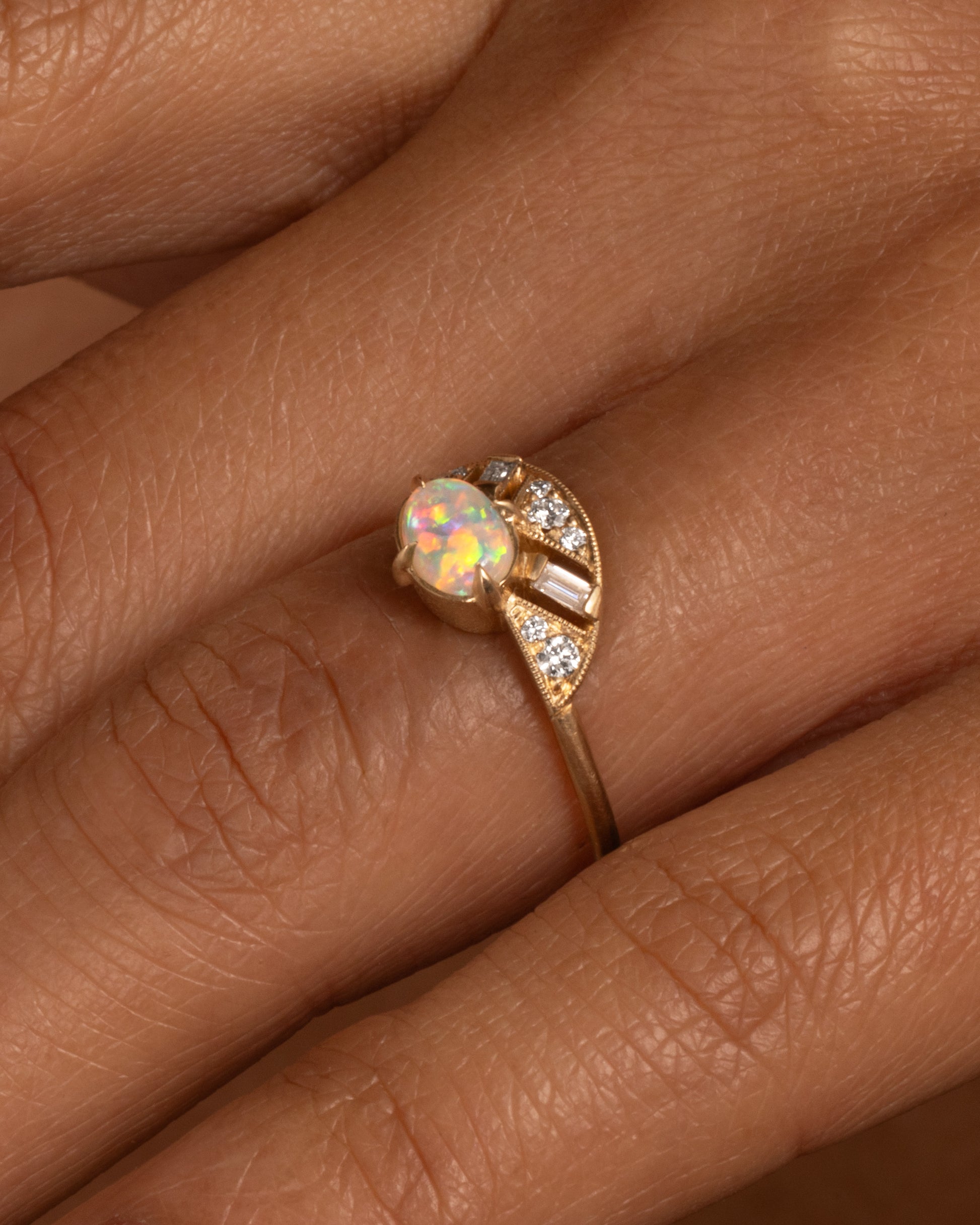 A yellow gold ring with a prong set opal and a diamond studded halo, shown on a hand.