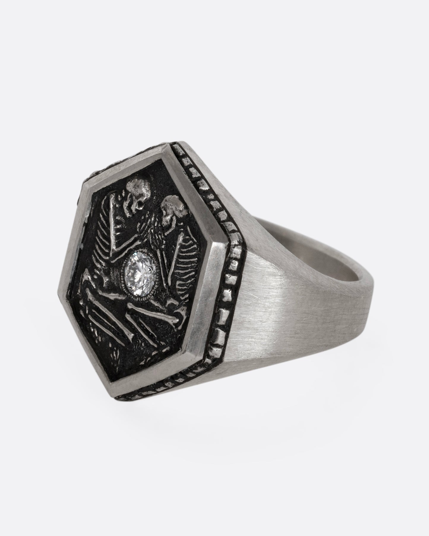 Sterling silver signet ring with two skeletons and a diamond at the center. Shown from the side.