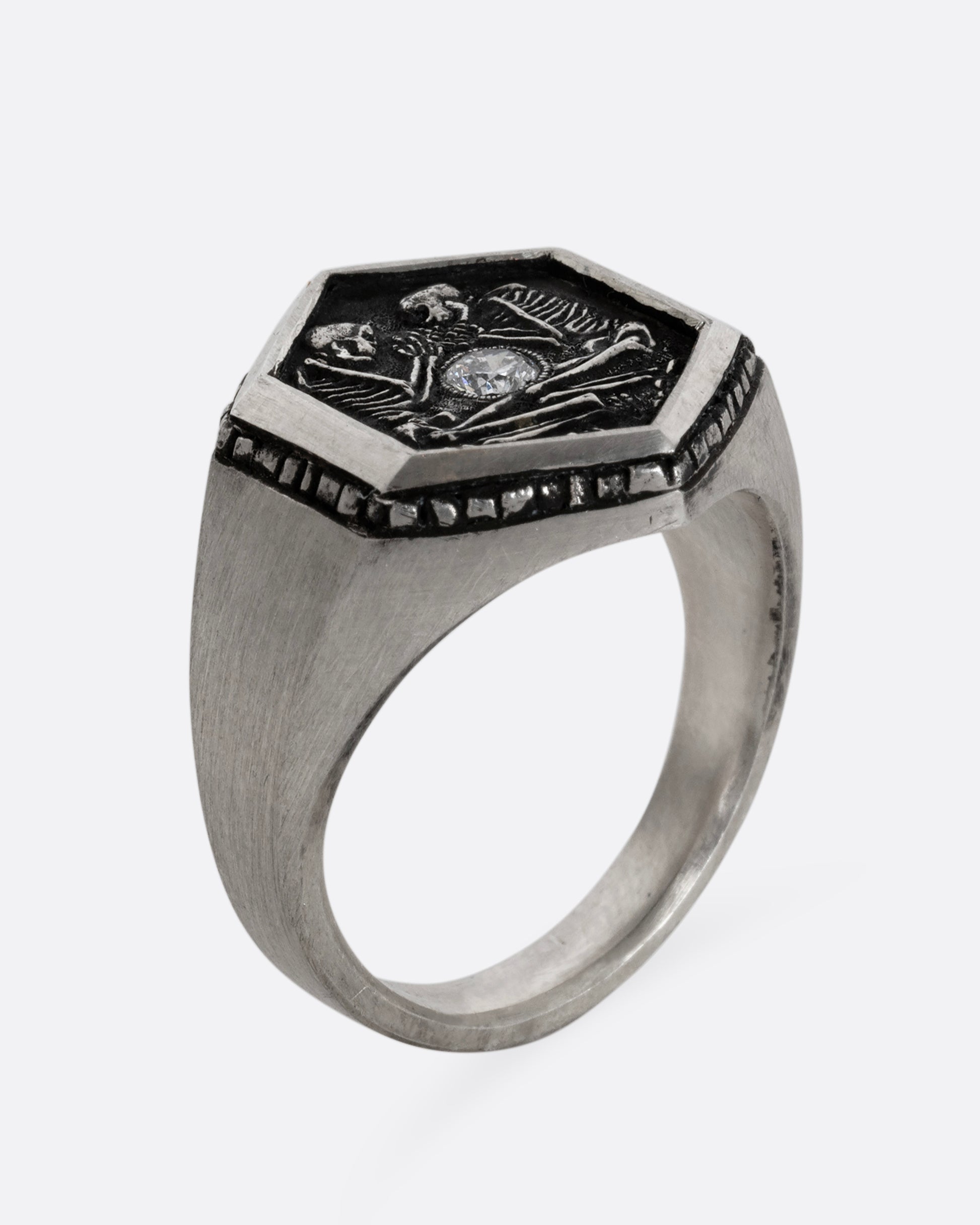 Sterling silver signet ring with two skeletons and a diamond at the center. Shown standing up from the side.