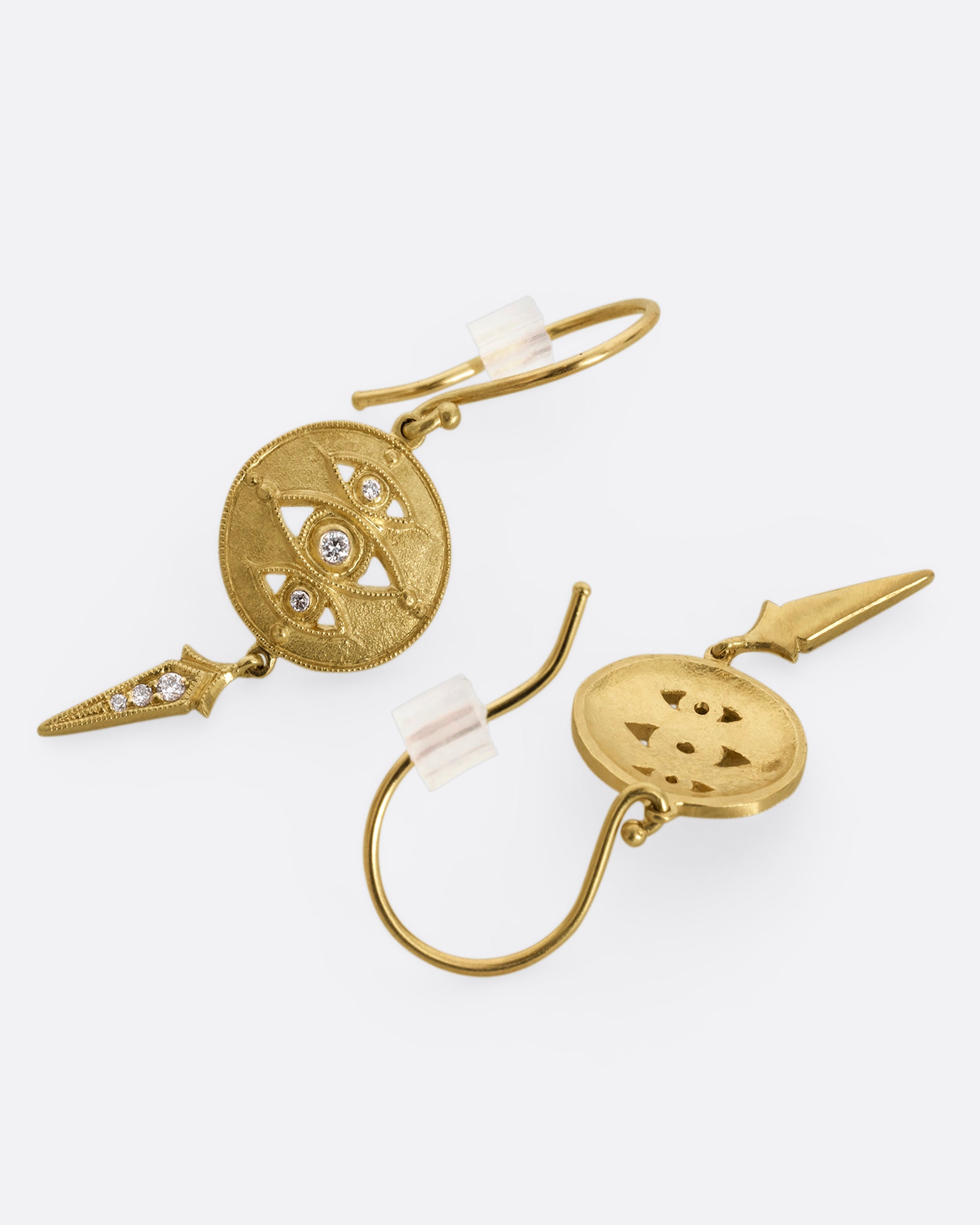 A pair of yellow gold drop earrings with diamond eyes on a disk and a spear shaped dangle. Shown laying flat.