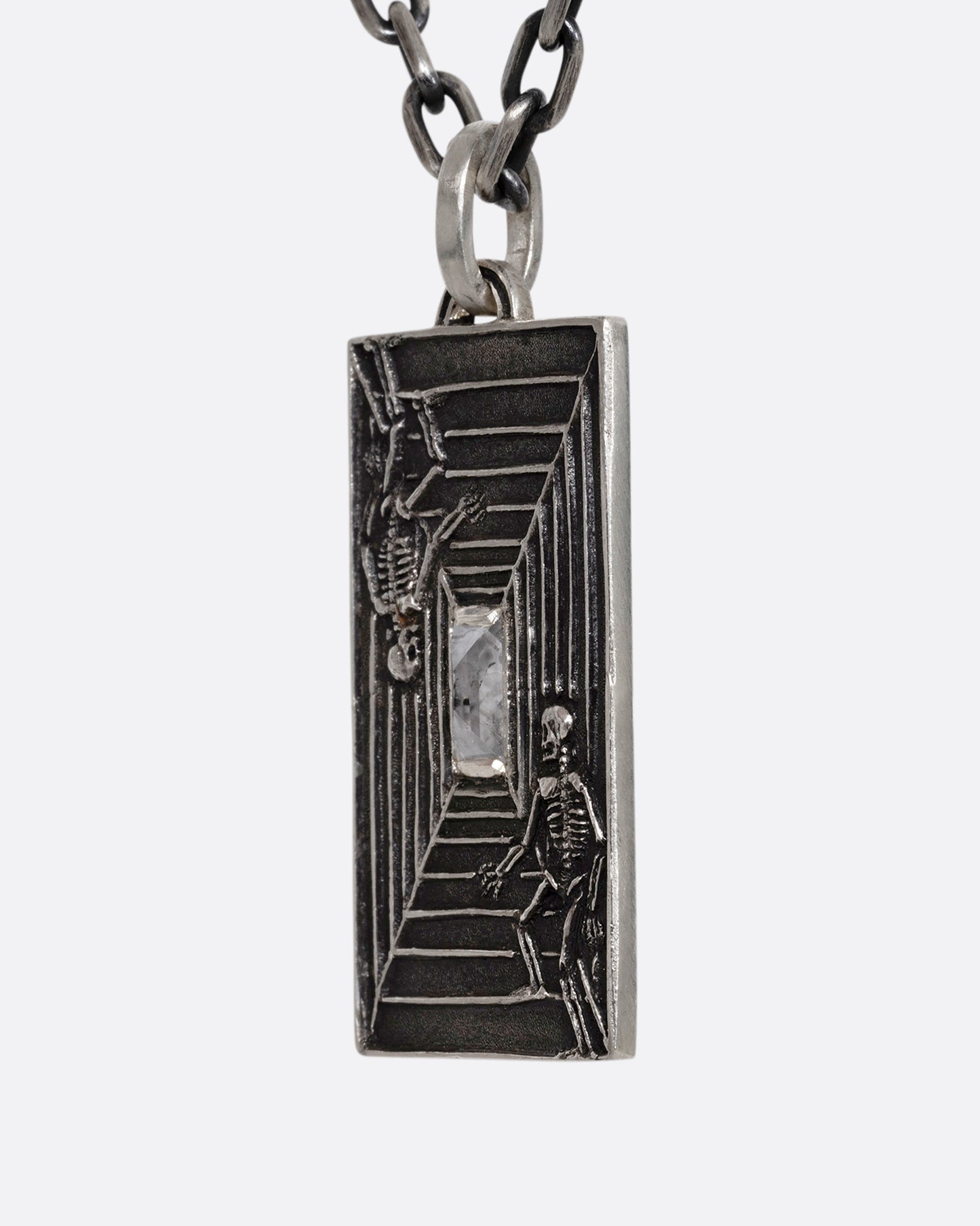 A sterling silver necklace with a rectangular pendant depicting two skeletons and a diamond at the center. Shown from the side.