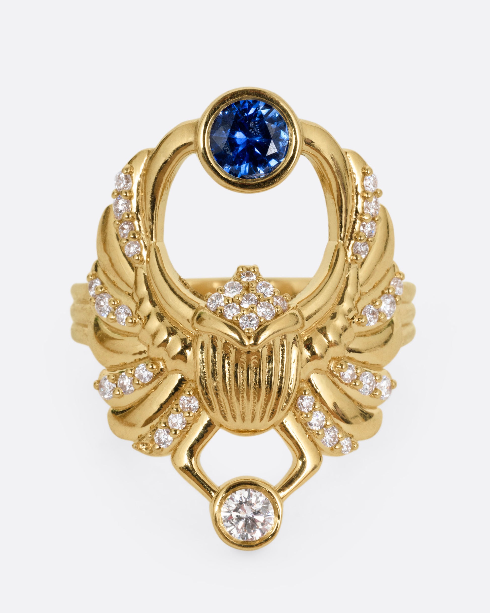 A yellow gold scarab ring with accents of white diamonds and a single blue sapphire between its fanned wings. Shown from the front.