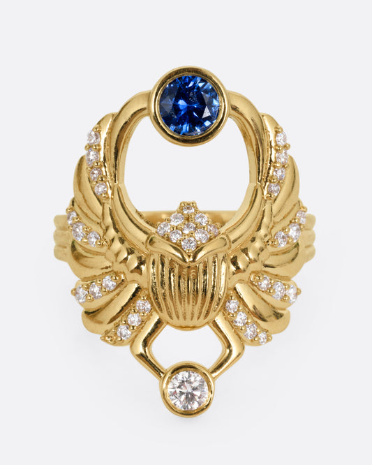 A yellow gold scarab ring with accents of white diamonds and a single blue sapphire between its fanned wings. Shown from the front.