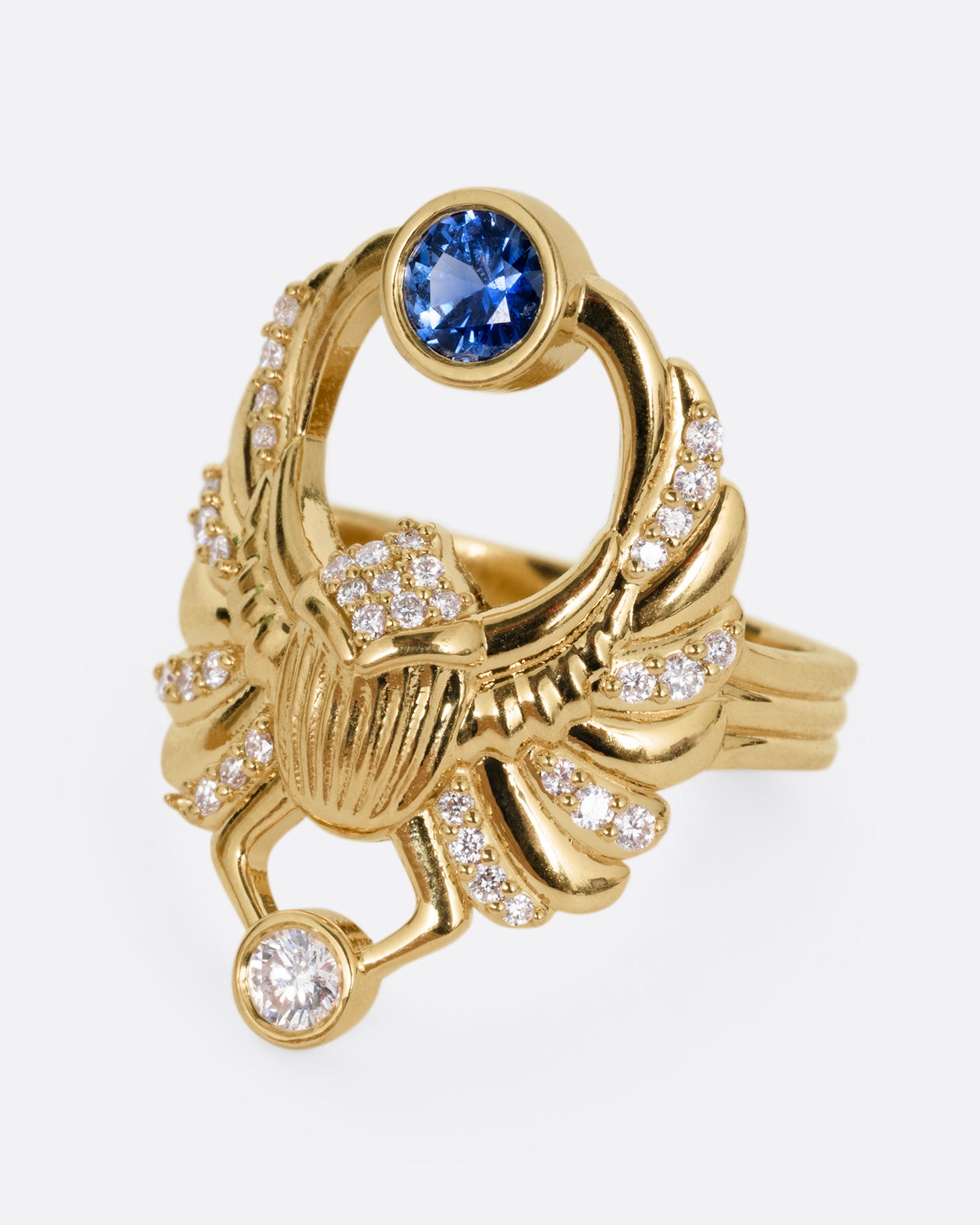 A yellow gold scarab ring with accents of white diamonds and a single blue sapphire between its fanned wings. Shown from the side.