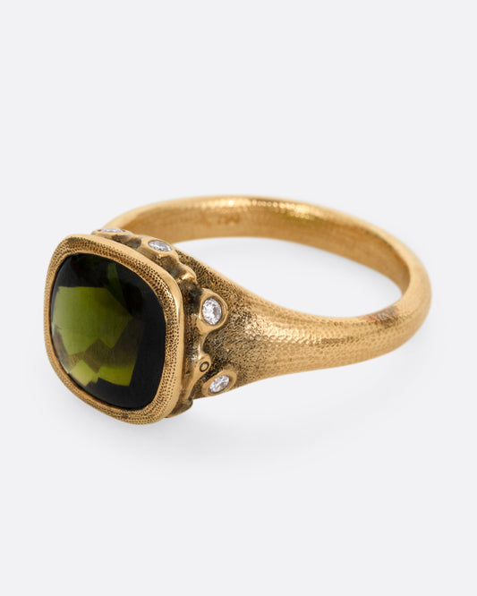 A yellow gold ring with a deep green tourmaline and white diamonds accents around its rounded edges. Shown from the side.