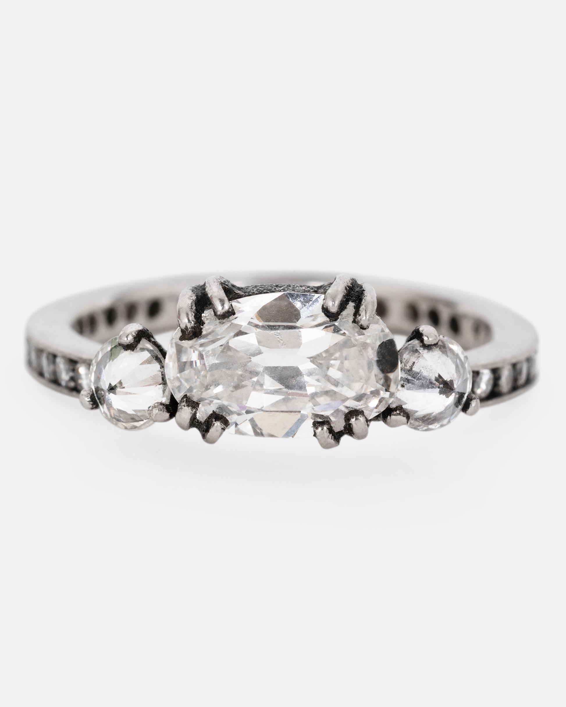 A platinum ring with a trio of diamonds, set upon a contrasting darkened channel of bead set diamonds. The elongated cushion cut center diamond is accented by two reverse set brilliant cut diamonds. Shown from the front. 