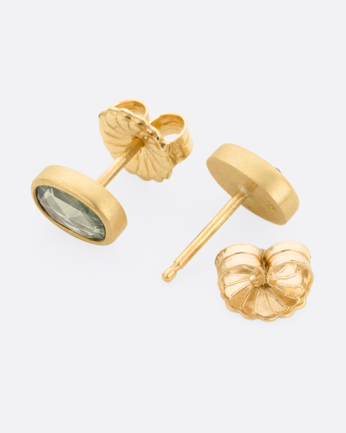 A pair of yellow gold stud earrings with pale green sapphires, shown lying on their sides, with one earring open.