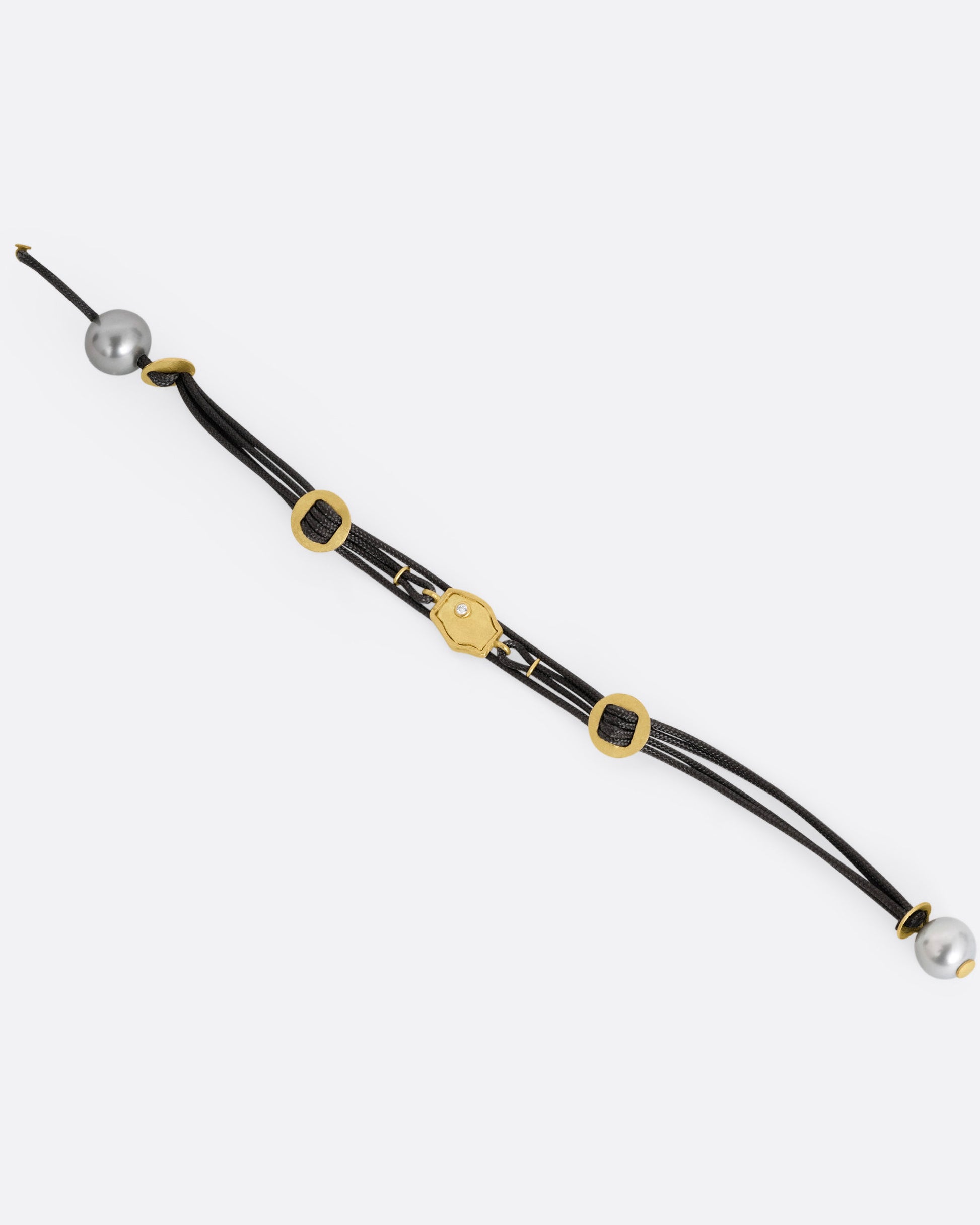 A  black cord bracelet with a diamond studded tag flanked by two gold slides and a pearl closure. Shown open from the front.
