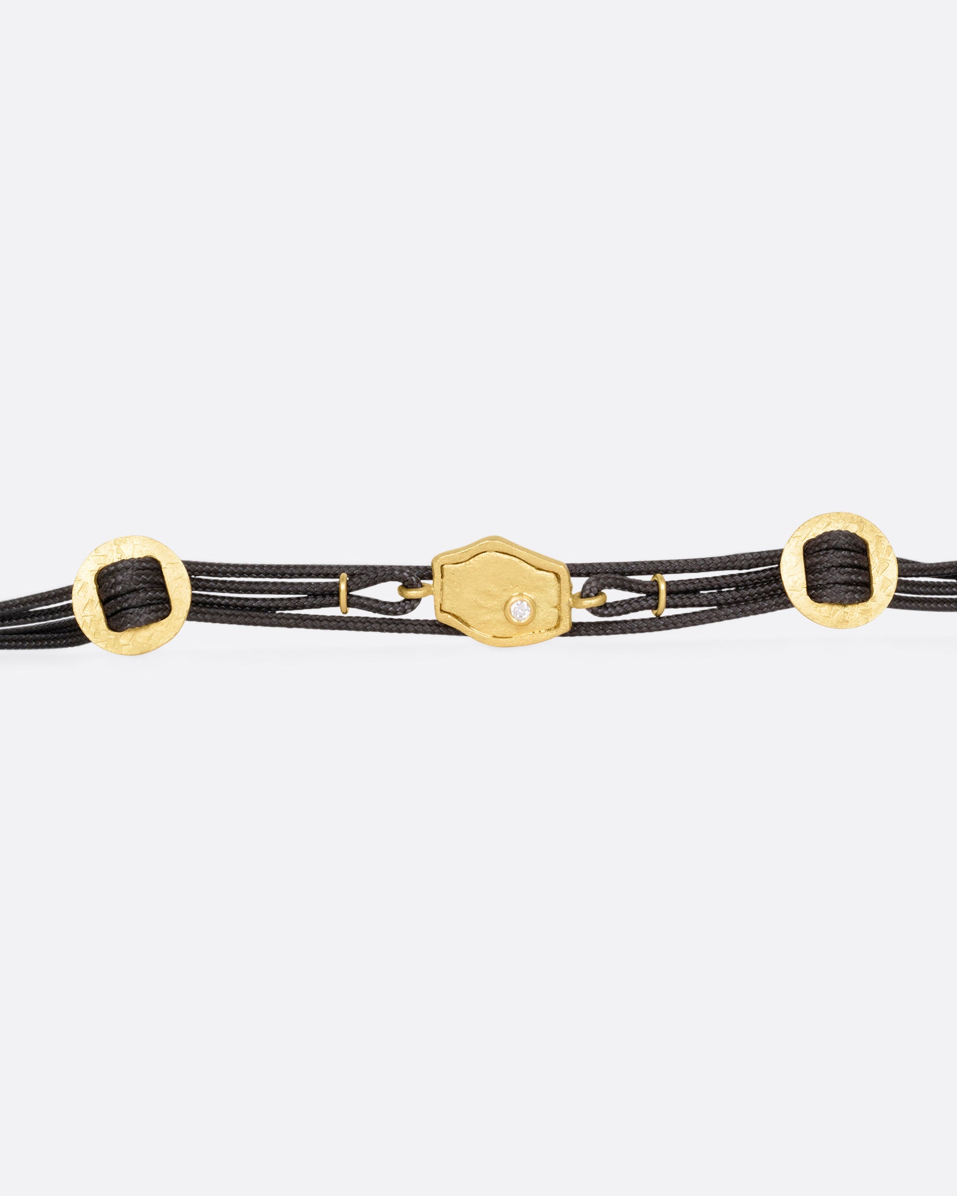 A  black cord bracelet with a diamond studded tag flanked by two gold slides and a pearl closure. Shown open from the front.