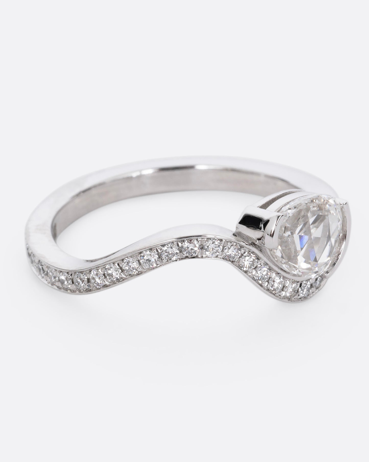 A diamond studded platinum ring with a wavy silhouette and a pear shaped rose cut diamond, shown from the side. 