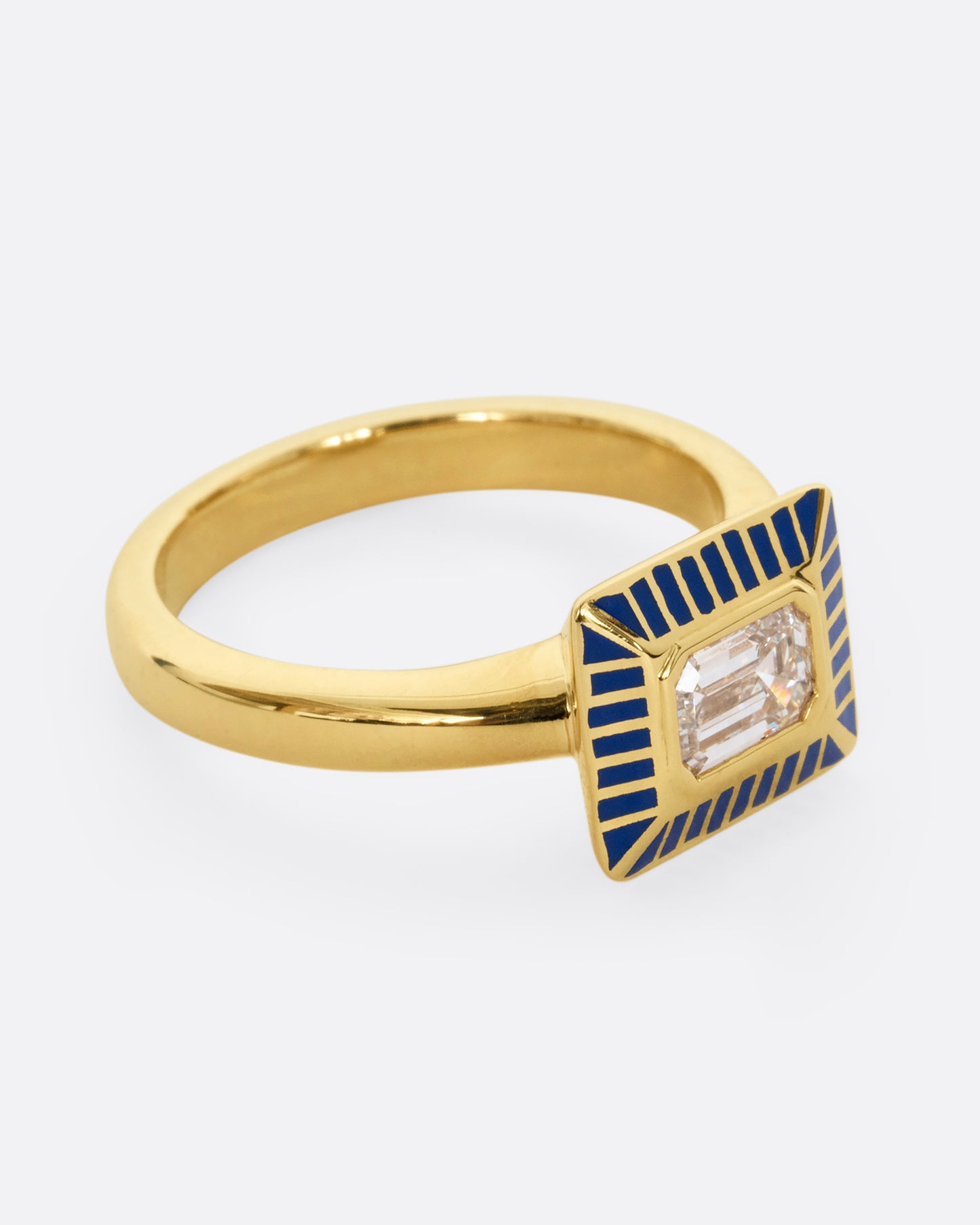 A yellow gold ring with an emerald cut diamond in a blue enamel striped rectangular setting. Shown from the side.