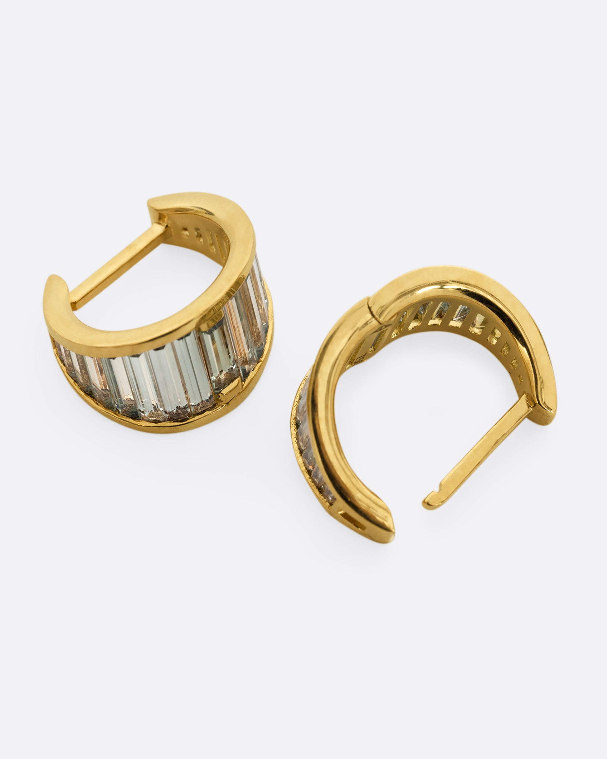 A pair of yellow gold huggie hoops with teal sapphire baguettes. Shown laying flat.