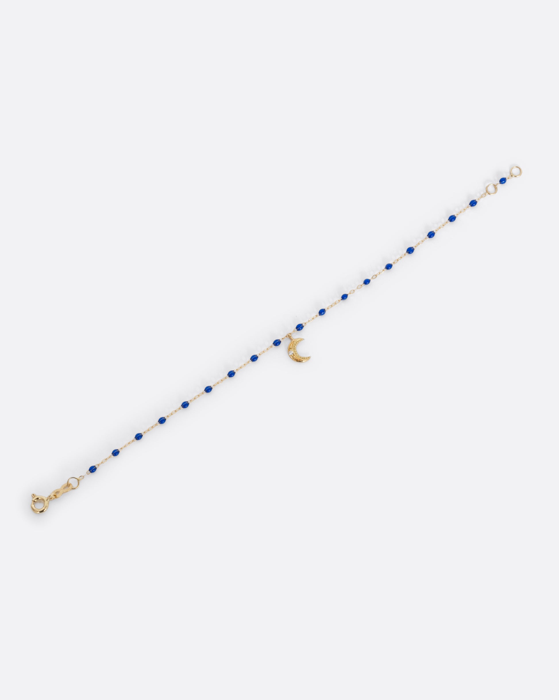 A yellow gold cable chain bracelet with blue resin droplets and a crescent moon charm. A single white diamond is set in the center of the crescent moon. Shown unclasped from the front.