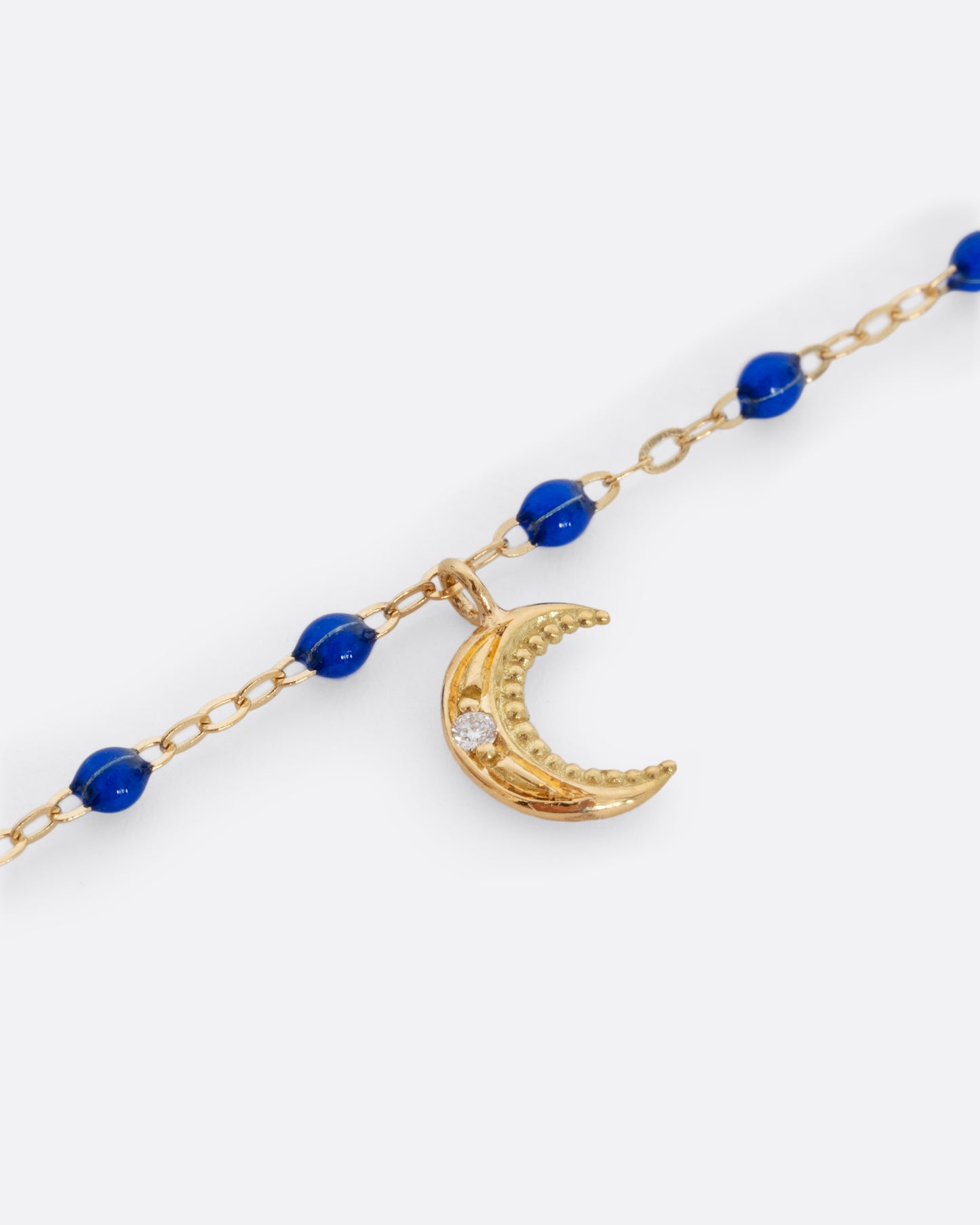 A close up shot of a yellow gold cable chain bracelet with blue resin droplets and a crescent moon charm. A single white diamond is set in the center of the crescent moon. Shown from the front.