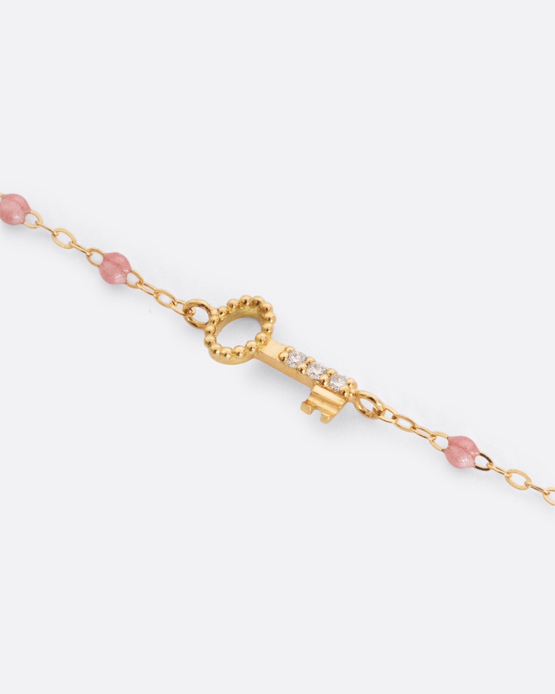 A close up shot of a yellow gold cable chain bracelet with droplets of pink resin and a yellow gold key charm. Three white diamonds are set on the key charm. Shown from the front.