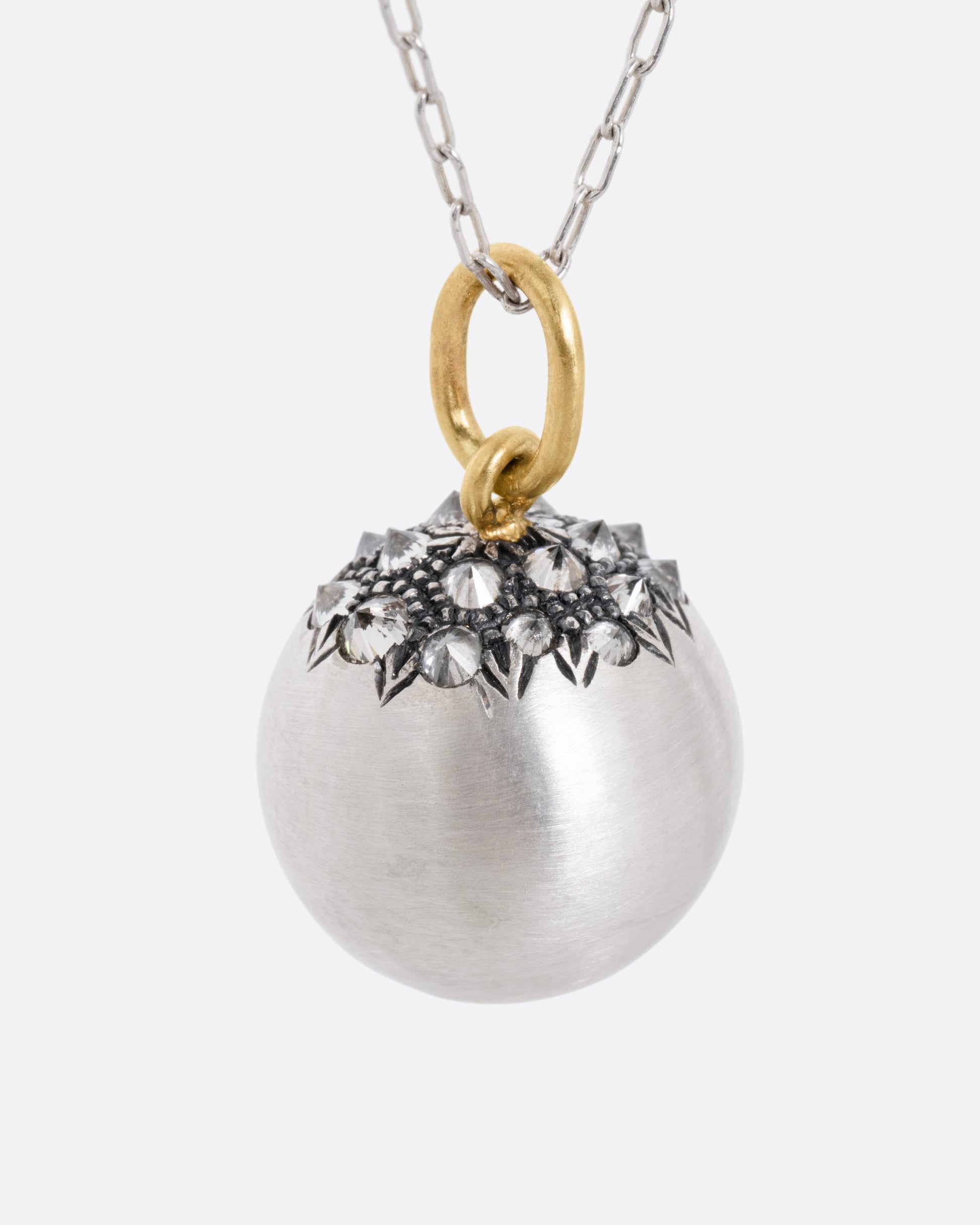 A silver cable chain necklace with a sizable sterling orb pendant peppered with grey reverse set diamonds and a yellow gold bail, shown from the side.
