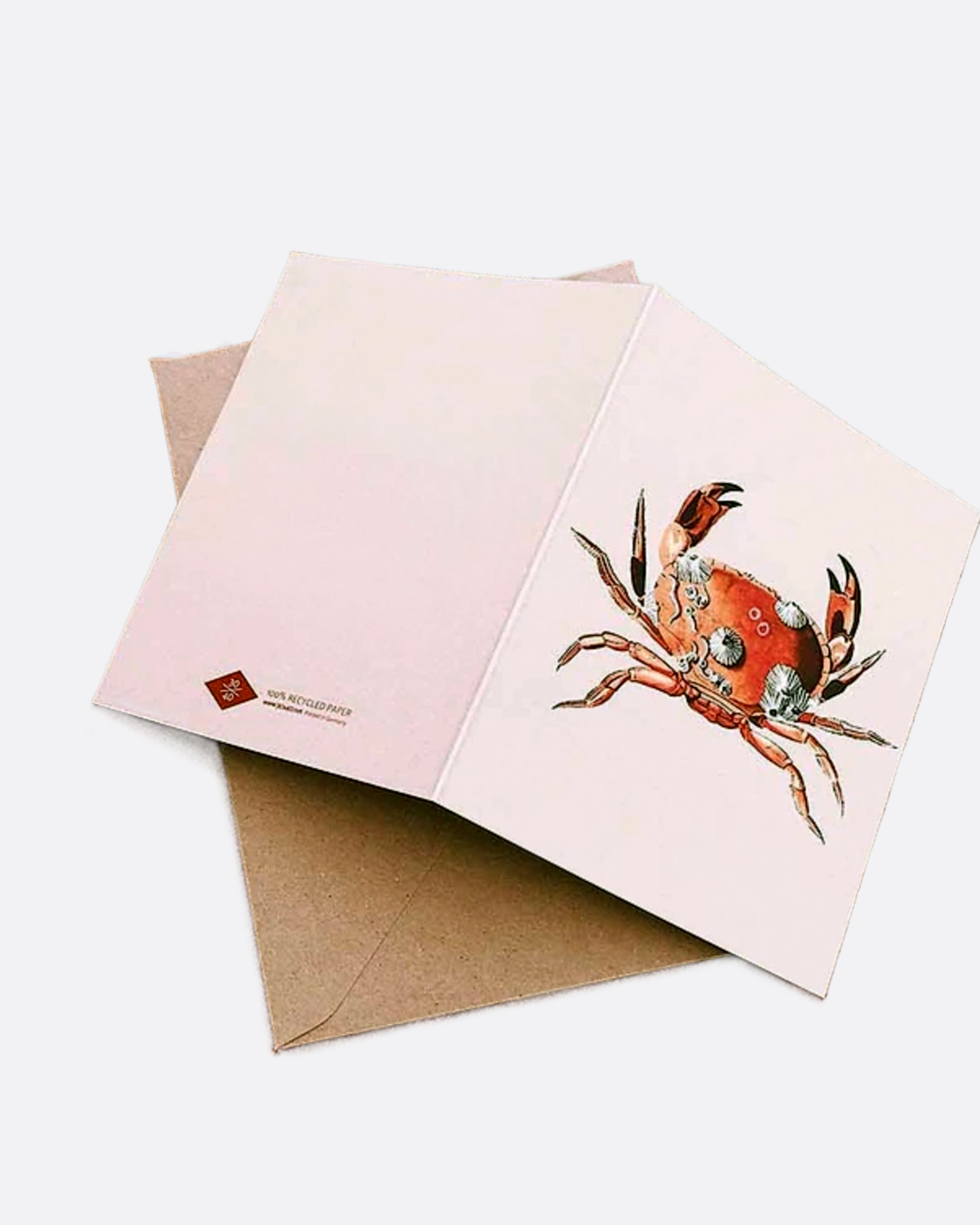 A light pink paper greeting card with red crab covered in barnacles on the front, a blank white interior, and a kraft paper envelope.