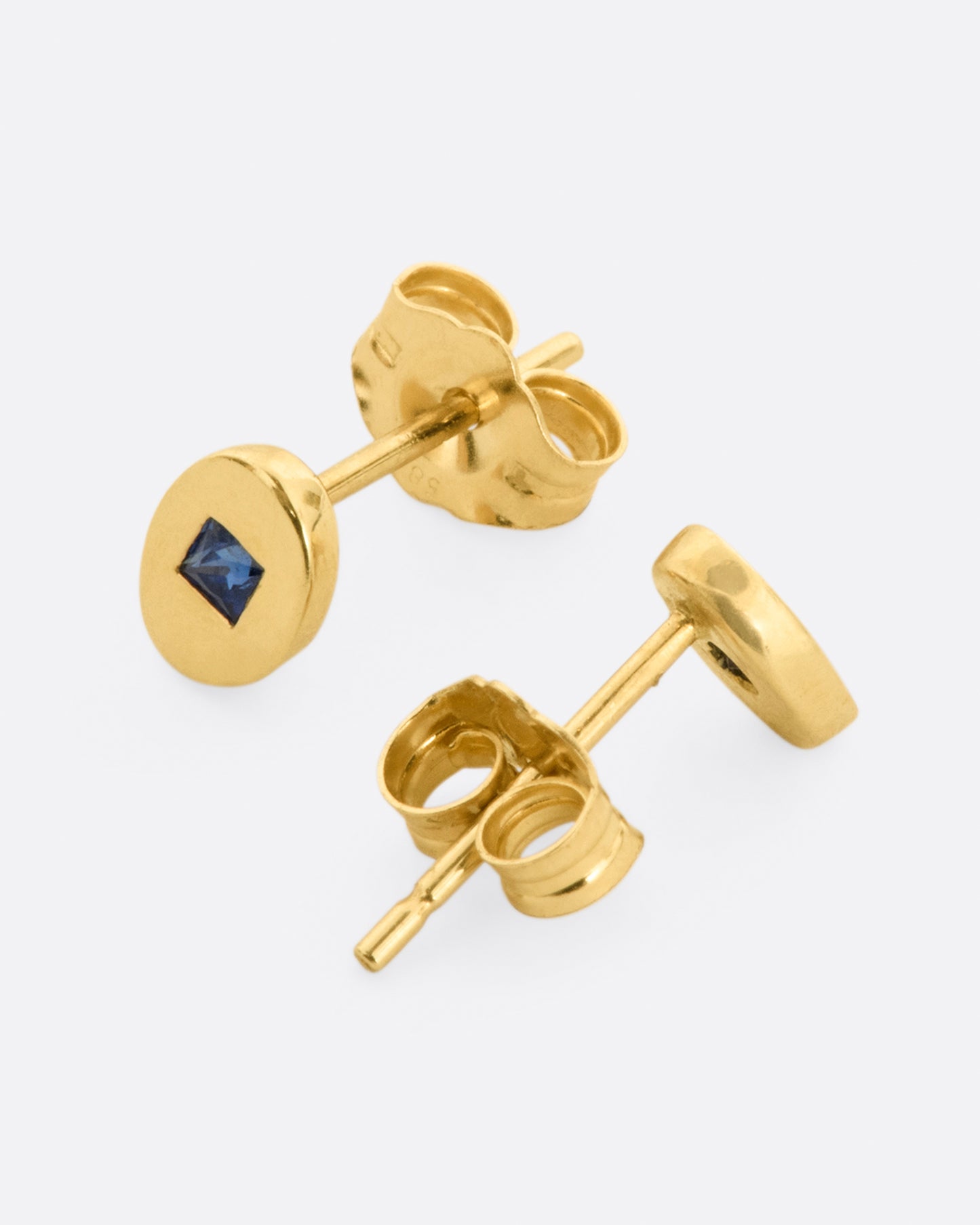 A pair of oval yellow gold stud earrings, each with a princess cut blue sapphire at its center. Shown from the side.