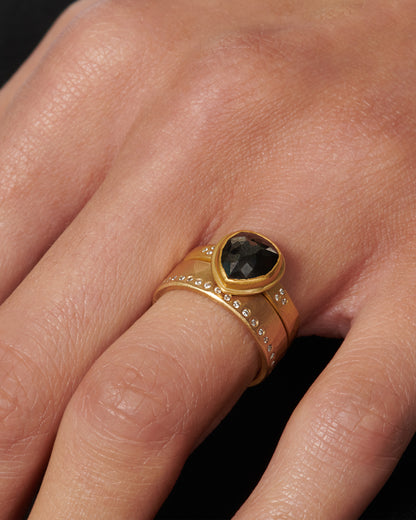 A hammered, matte gold band with a line of round diamonds all the way around. Shown stacked with a large teardrop shaped black diamond yellow gold ring on a hand.